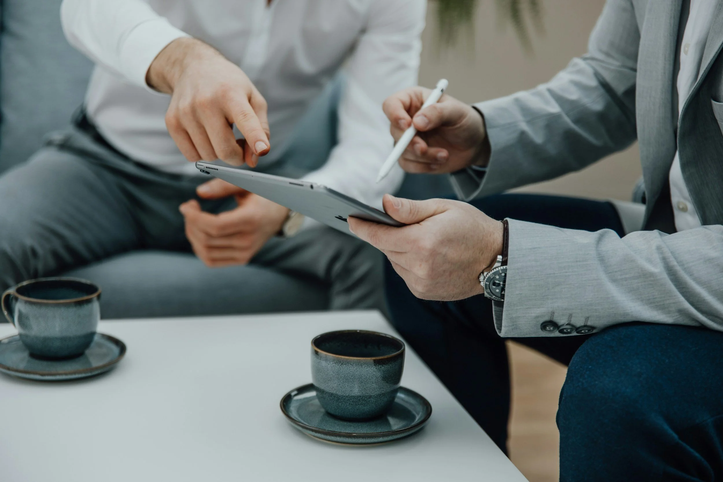 Two men in business attire consulting over a digital tablet with coffee cups on a table.