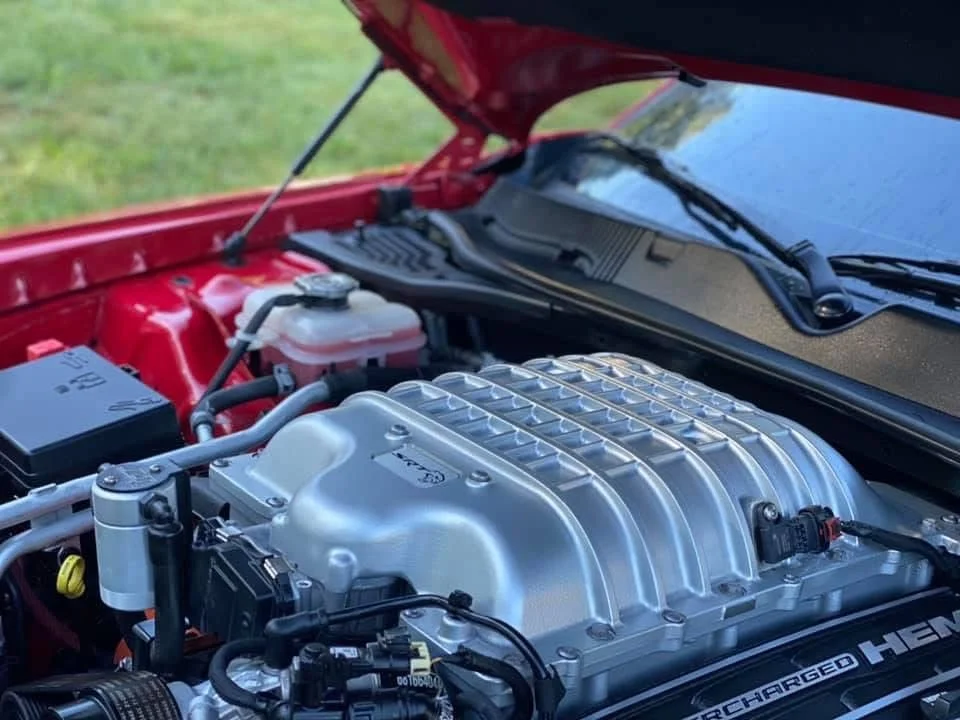 Close-up of a car engine, featuring a supercharger with a silver finish, mounted in a red engine bay, with various hoses, wires, and components visible.