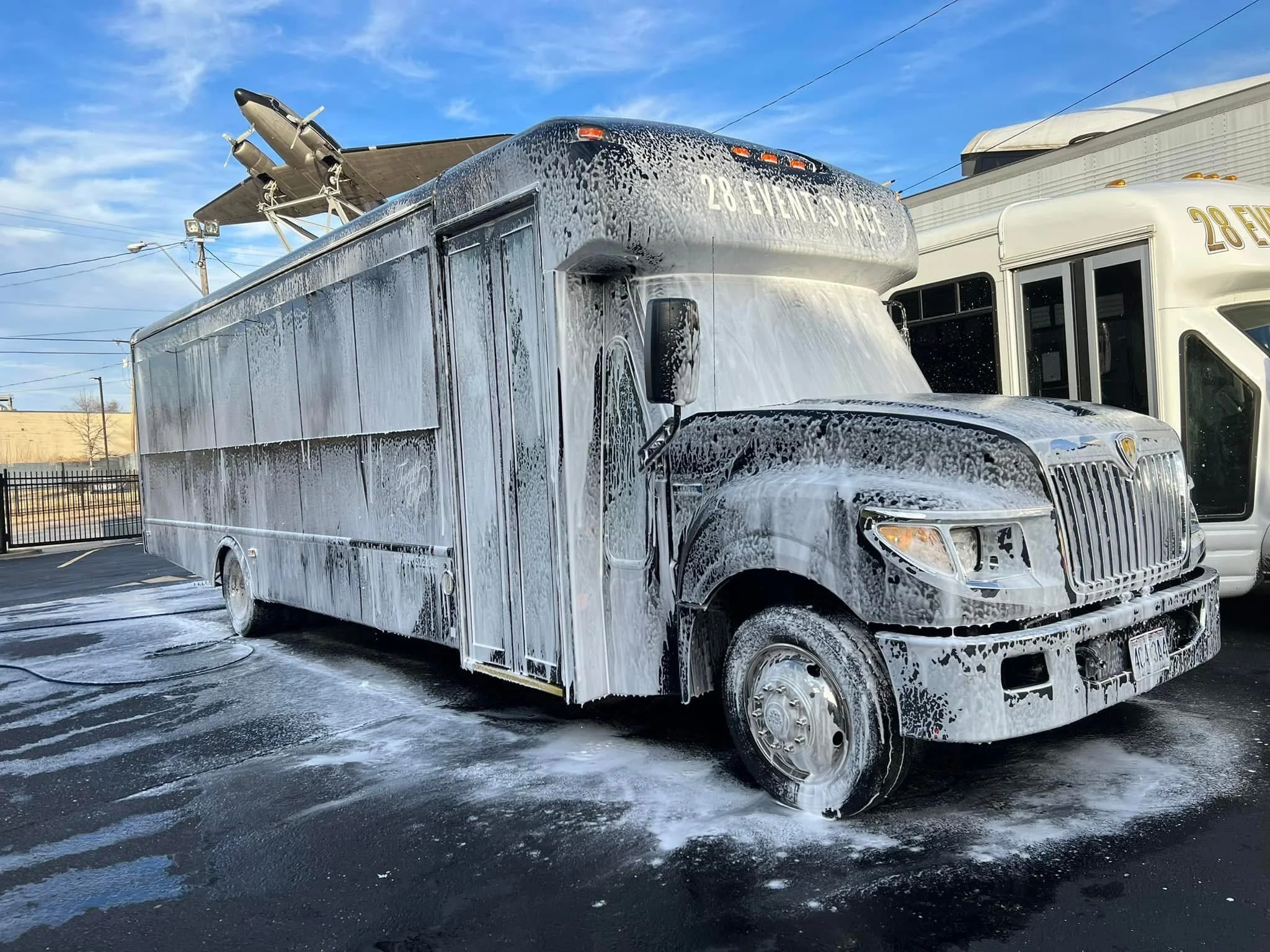 A foam-covered food truck with a soap and water spray hose on the ground in front of it, parked outdoors with another similar vehicle nearby.