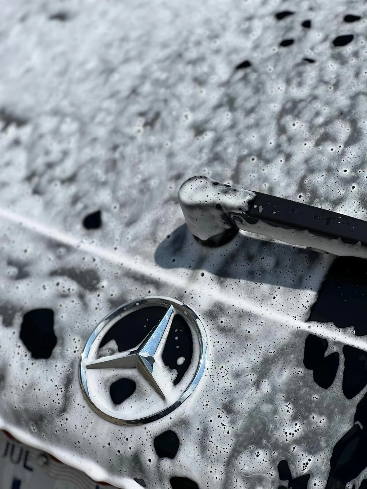 Mercedes-Benz logo on a car hood covered in soap suds during washing.