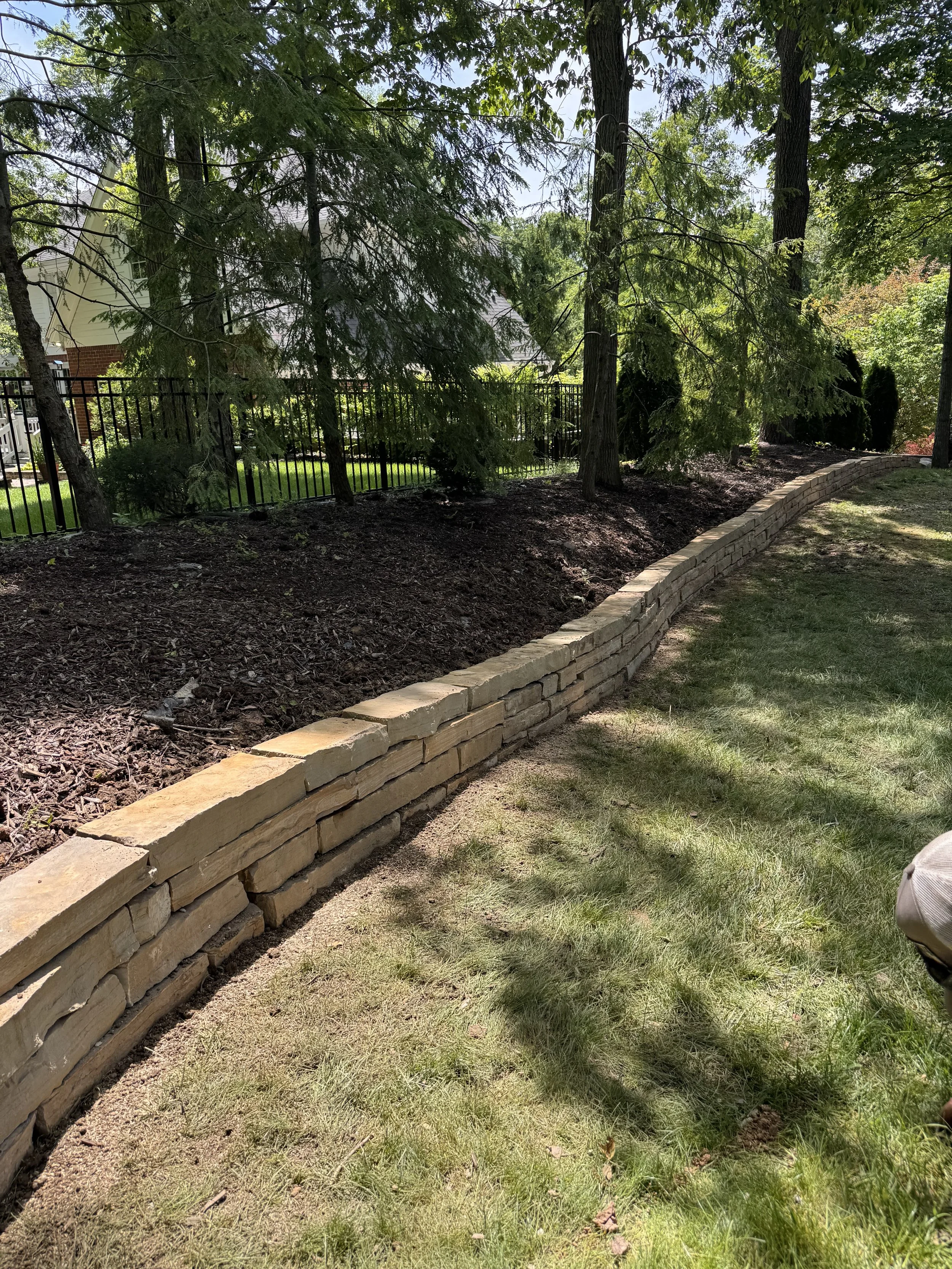 A landscaped backyard with a stone retaining wall, trees, and a black metal fence in front of a house with vinyl siding and a brick foundation.