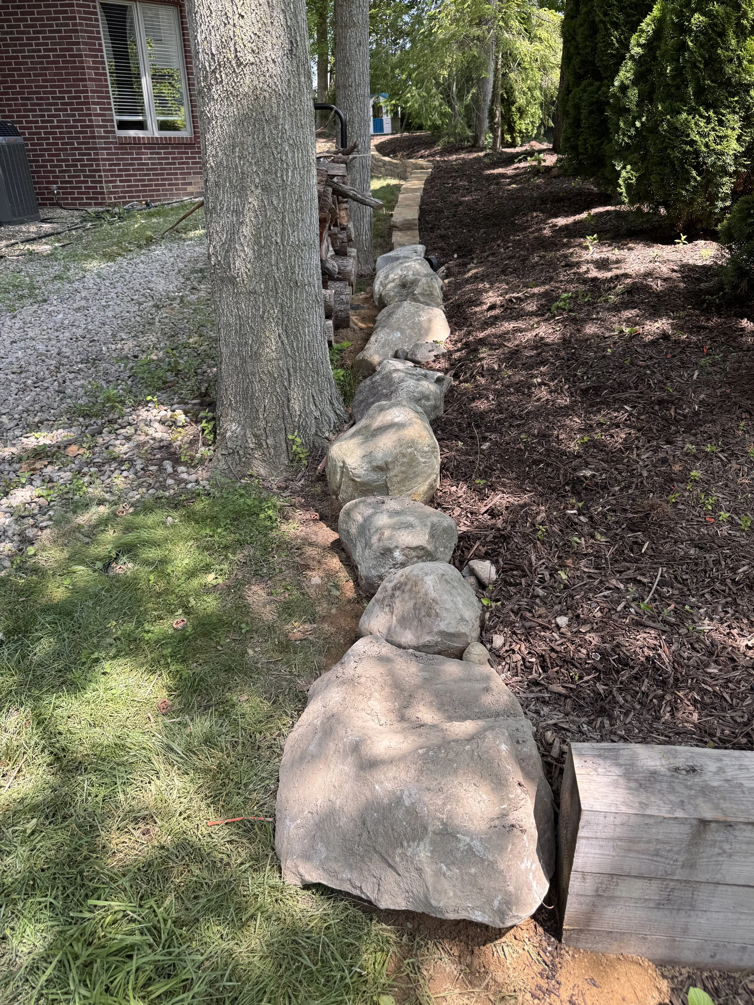 A line of large decorative rocks borders a newly landscaped garden bed with dark mulch along a house with red brick exterior and white-framed window. There are trees and a stone pathway in the background.