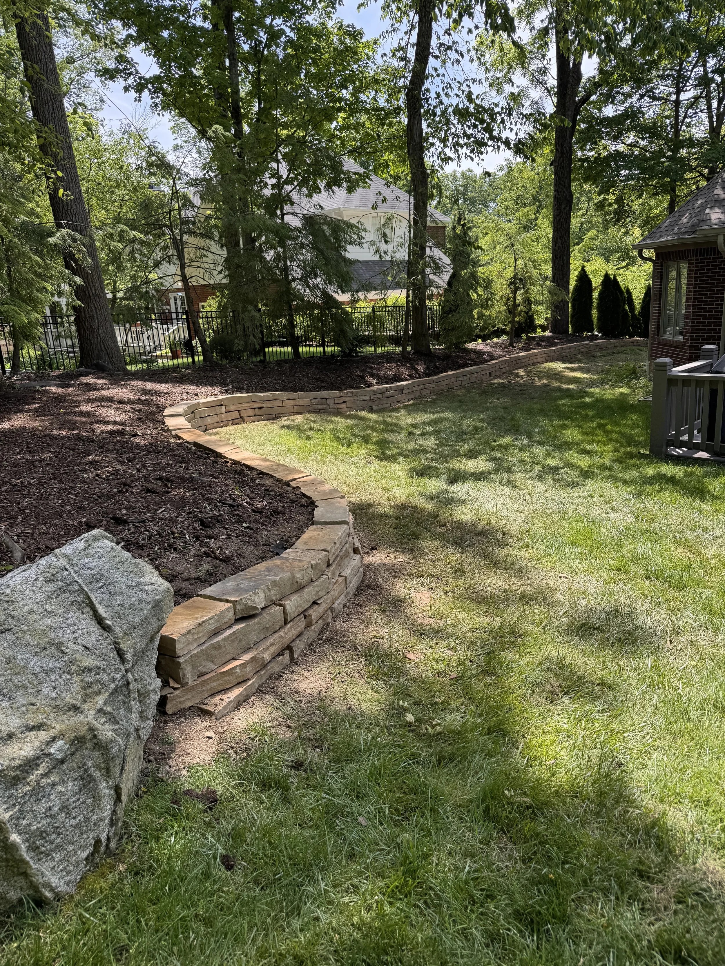 A landscaped backyard with a curved brick retaining wall, green grass, large trees, and a house in the background.