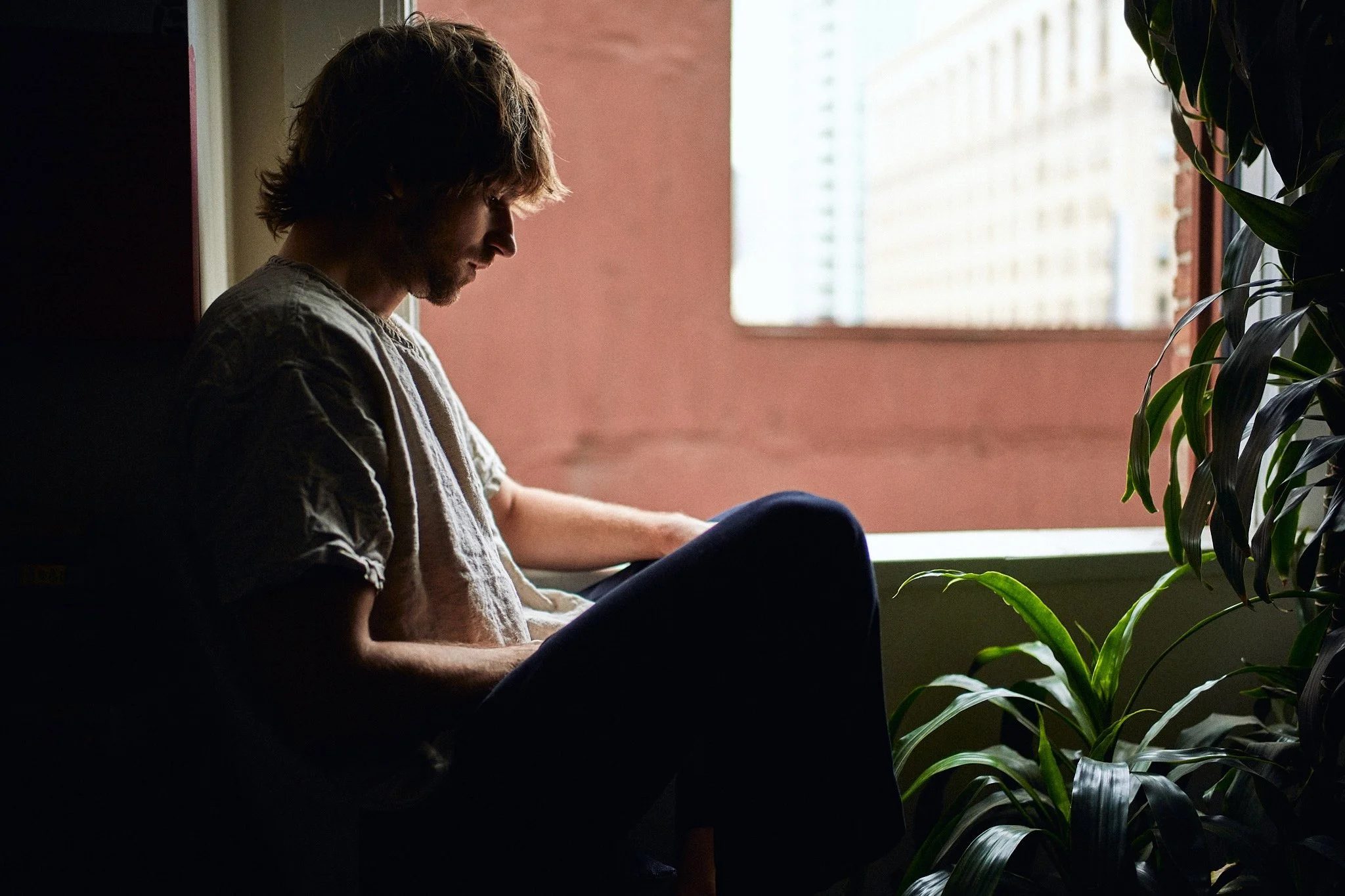 A young man with shaggy hair sitting on a dark chair near a window, looking down, with sunlight streaming in and greenery nearby.