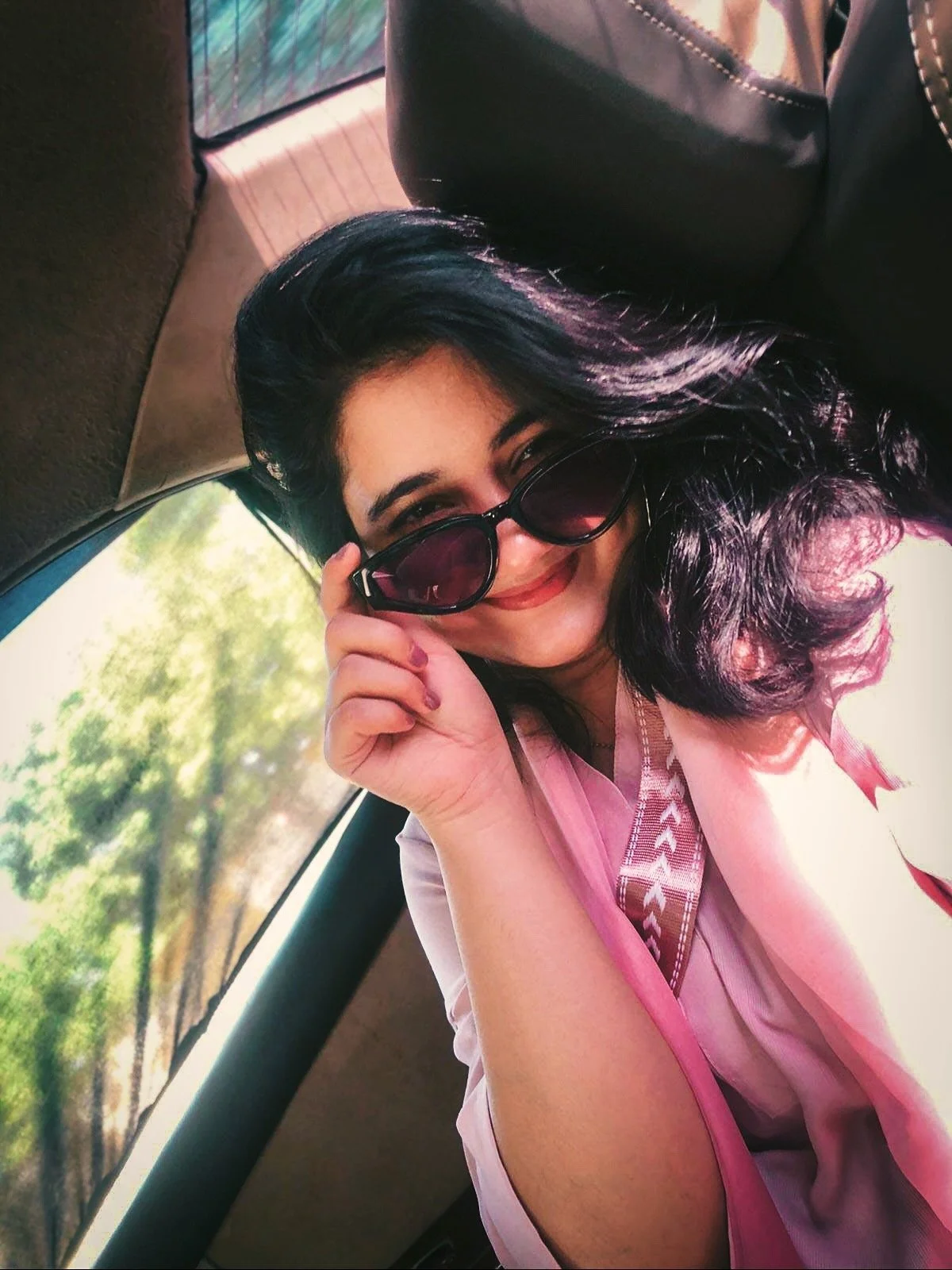 A woman with dark hair and sunglasses smiling and holding her glasses, sitting in a vehicle with a window showing trees outside.