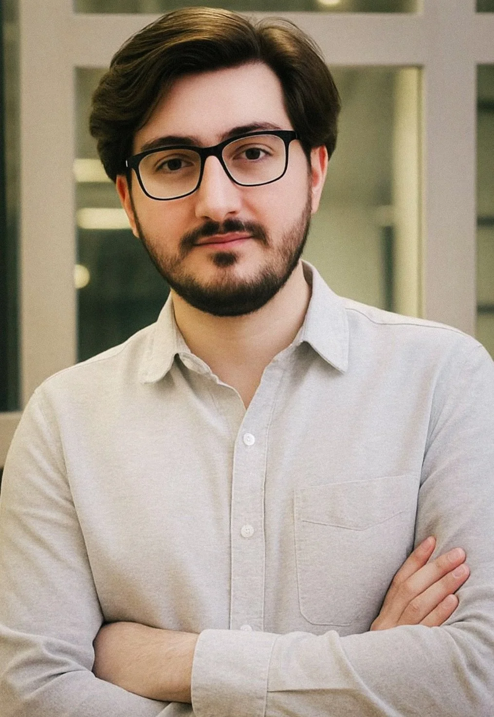 A young man with dark hair, glasses, and facial hair wearing a light-colored button-up shirt, standing with arms crossed indoors.