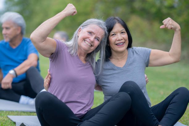 Two women sitting outdoors on a yoga mat, flexing their arms and smiling at the camera, happy with the results and mobility the in-home physical therapy provided them.