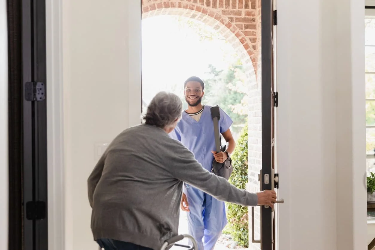 A senior woman  opens the front door to her in-home physical therapist in blue scrubs, ready to start her in-home physical therapy appointment in Boynton Beach, Florida.