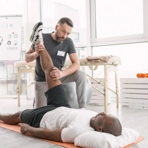 A physical therapist assisting a man with leg stretching exercises in a physical therapy clinic.
