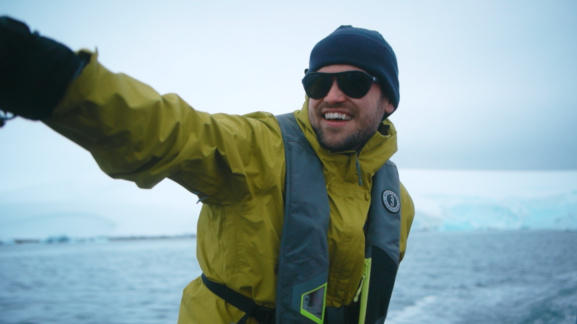 A smiling man wearing a yellow waterproof jacket, black beanie, dark sunglasses, and a life vest, taking a selfie on a boat with an ocean and ice in the background.