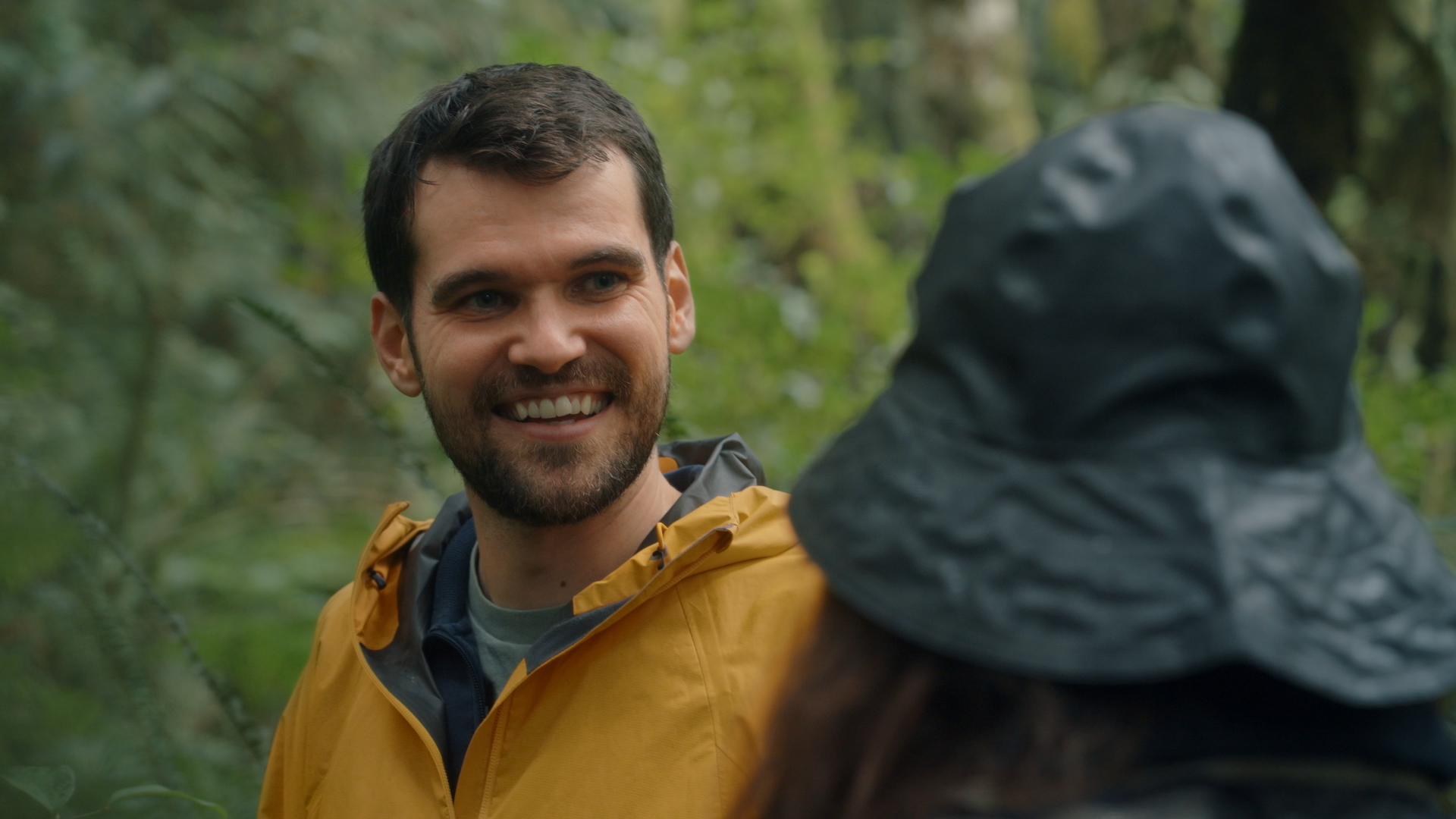 A smiling man in an orange rain jacket talking to a woman wearing a gray hat in a forest.