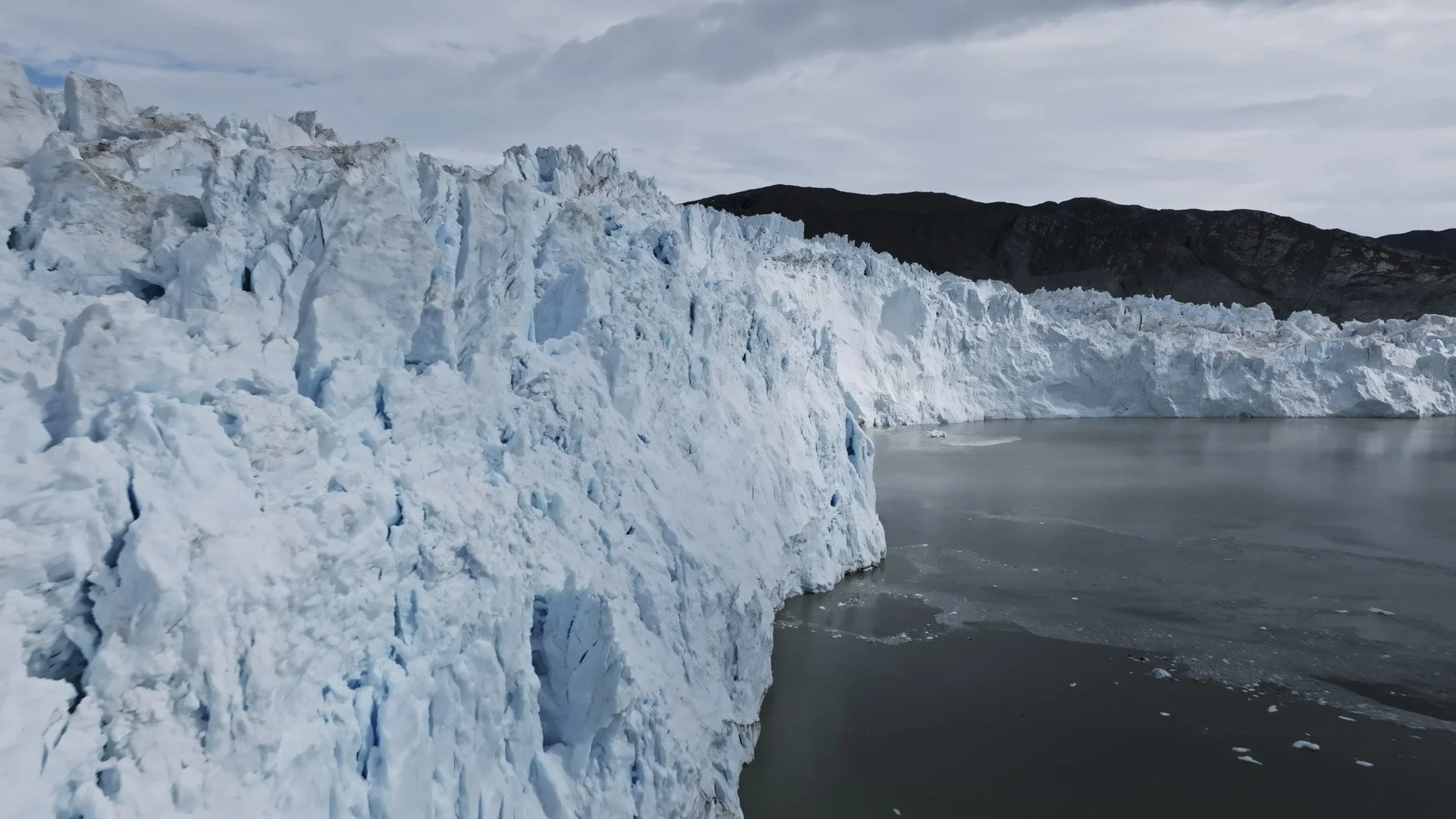 Large ice glacier meeting the sea with dark mountains in the background under a cloudy sky.