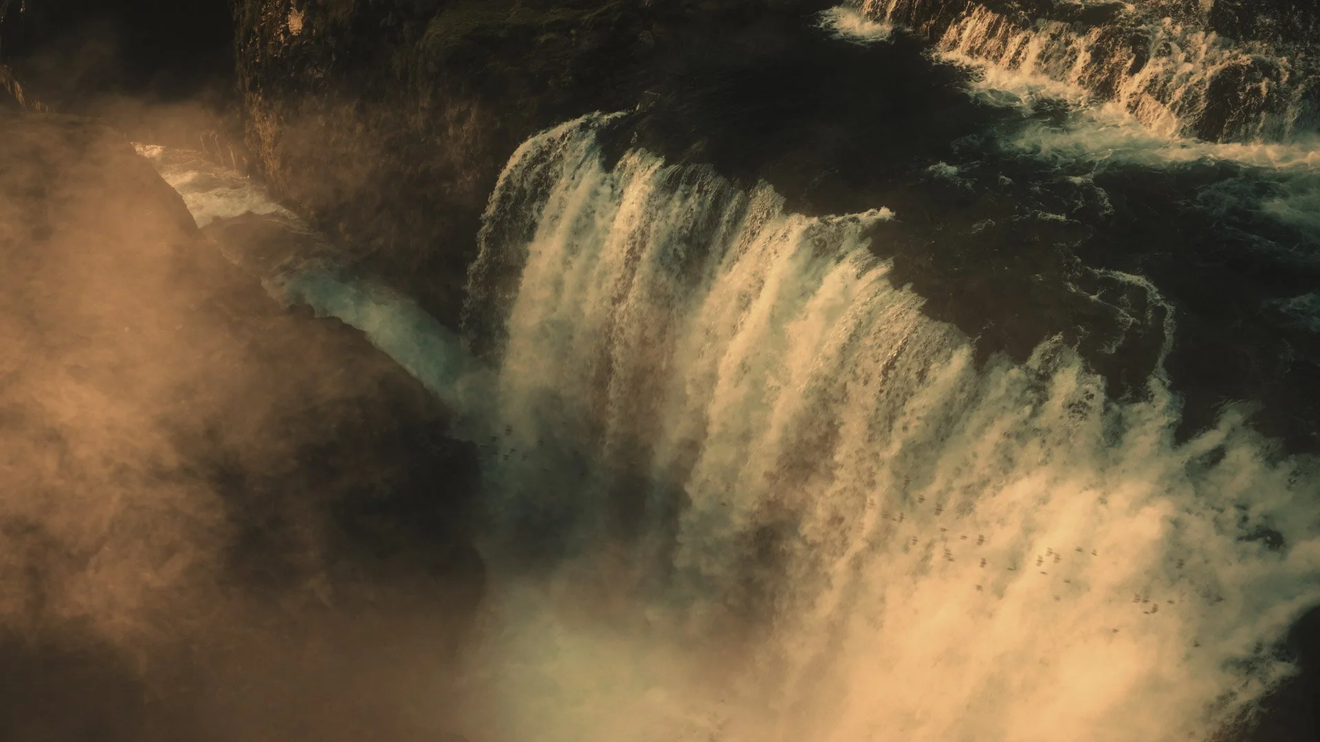 A large waterfall cascading over a dark rocky cliff with mist rising at the base, illuminated with a warm, golden light.