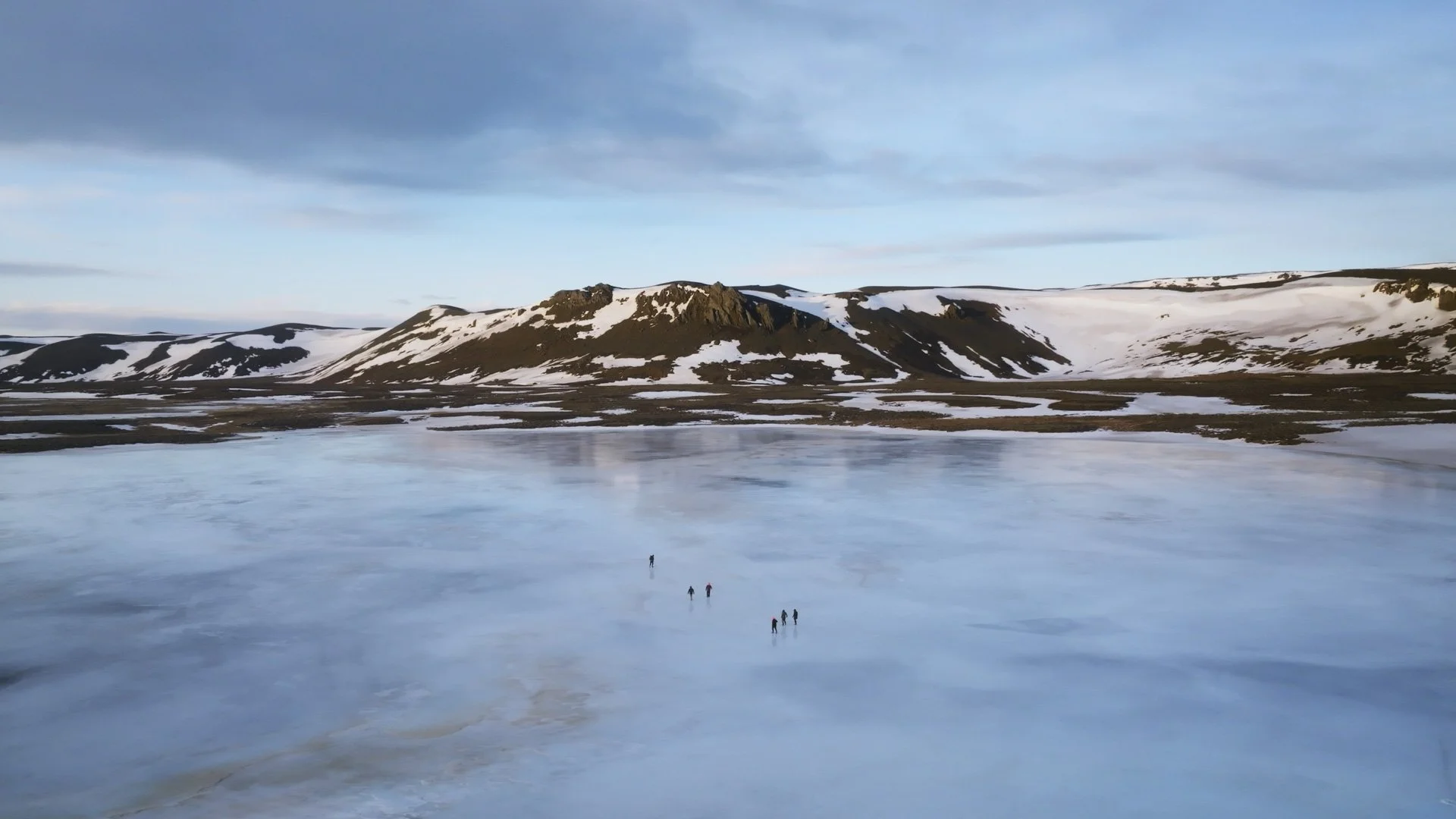A frozen lake with small groups of hikers walking across, surrounded by snow-covered mountains and a partly cloudy sky.