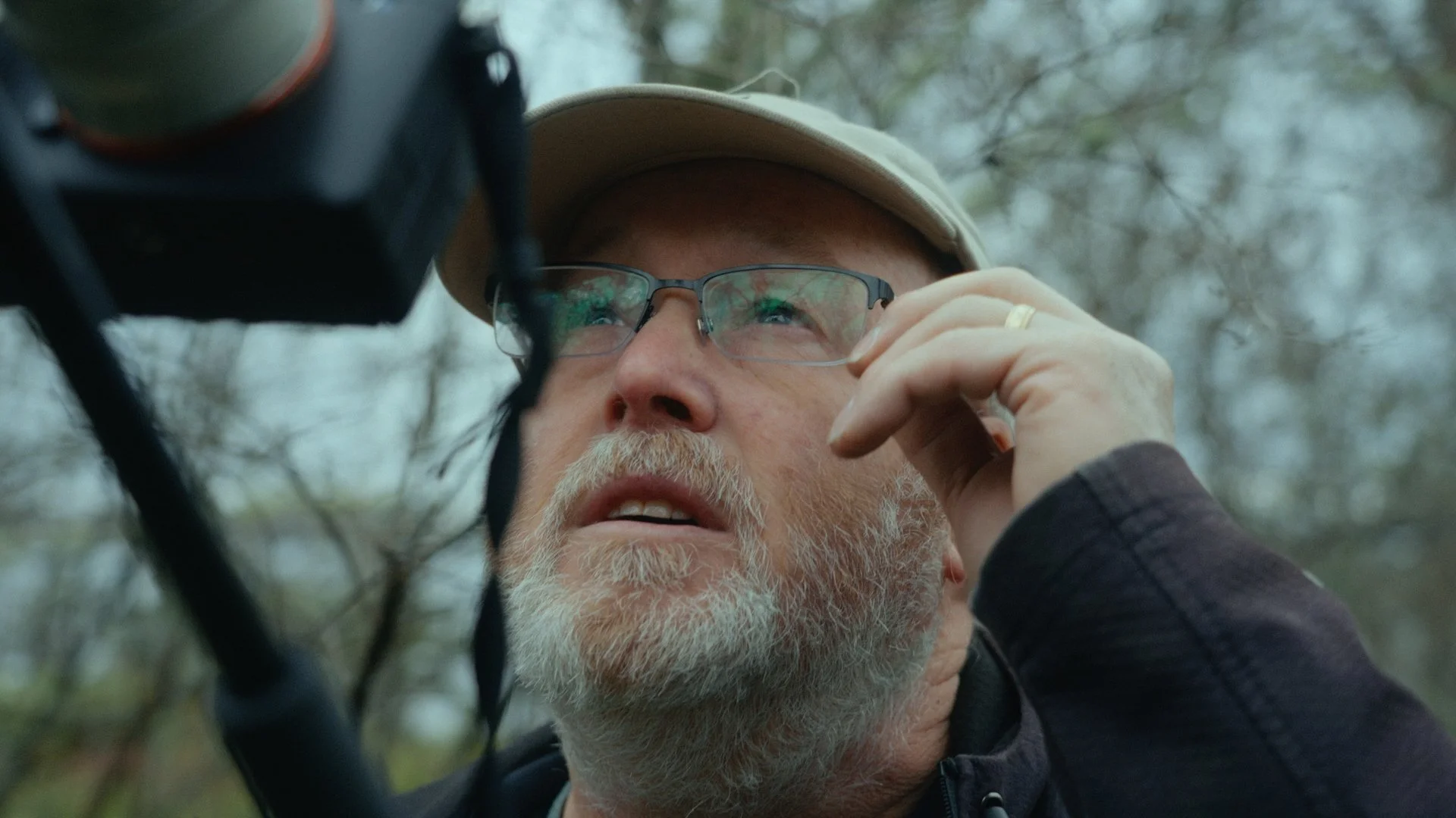 Close-up of an older man with a beard and glasses looking upward while adjusting his glasses outdoors.