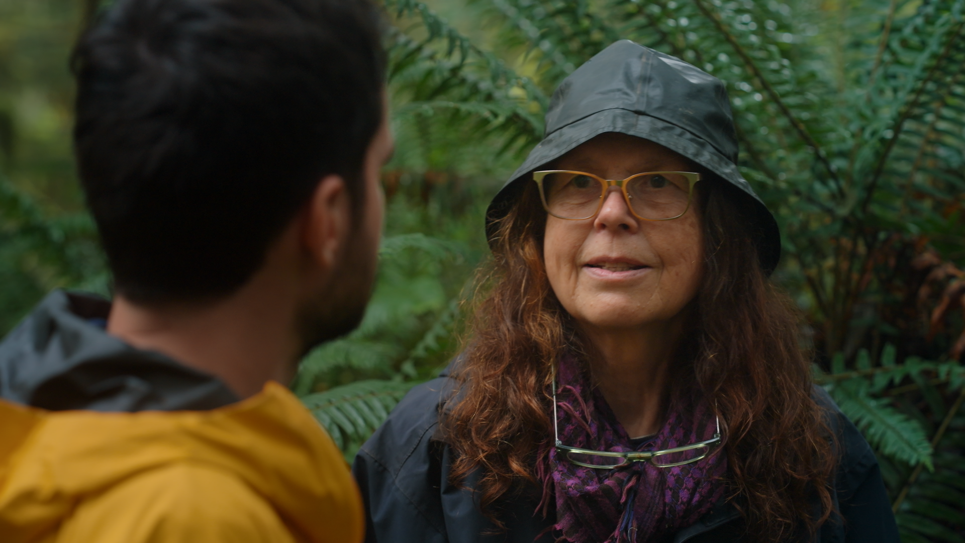 Two people, one woman and one man, engaged in conversation in a lush, green forest. The woman is wearing a black rain hat, glasses, and a scarf, and appears to be speaking, while the man, wearing a yellow jacket, is listening.