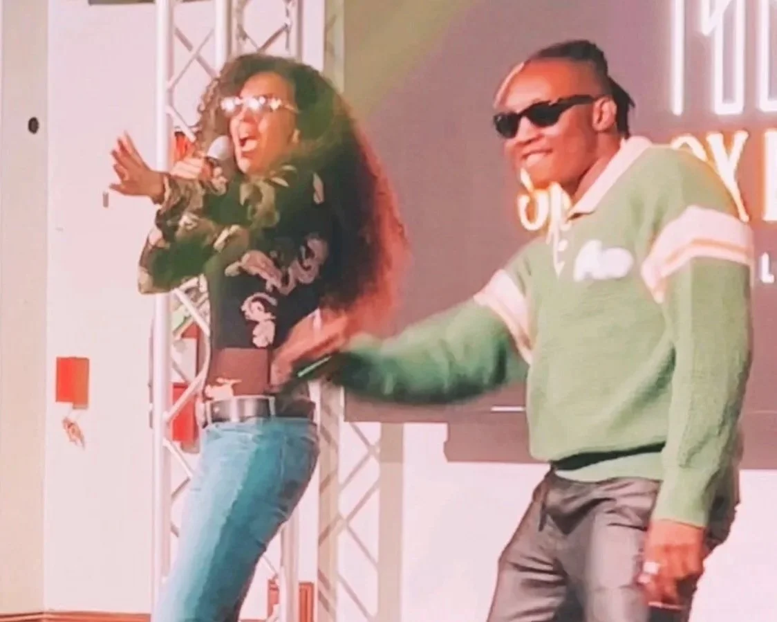 A woman with curly hair and sunglasses singing into a microphone, and a man with sunglasses smiling and wearing a green sweater, on stage with a backdrop.