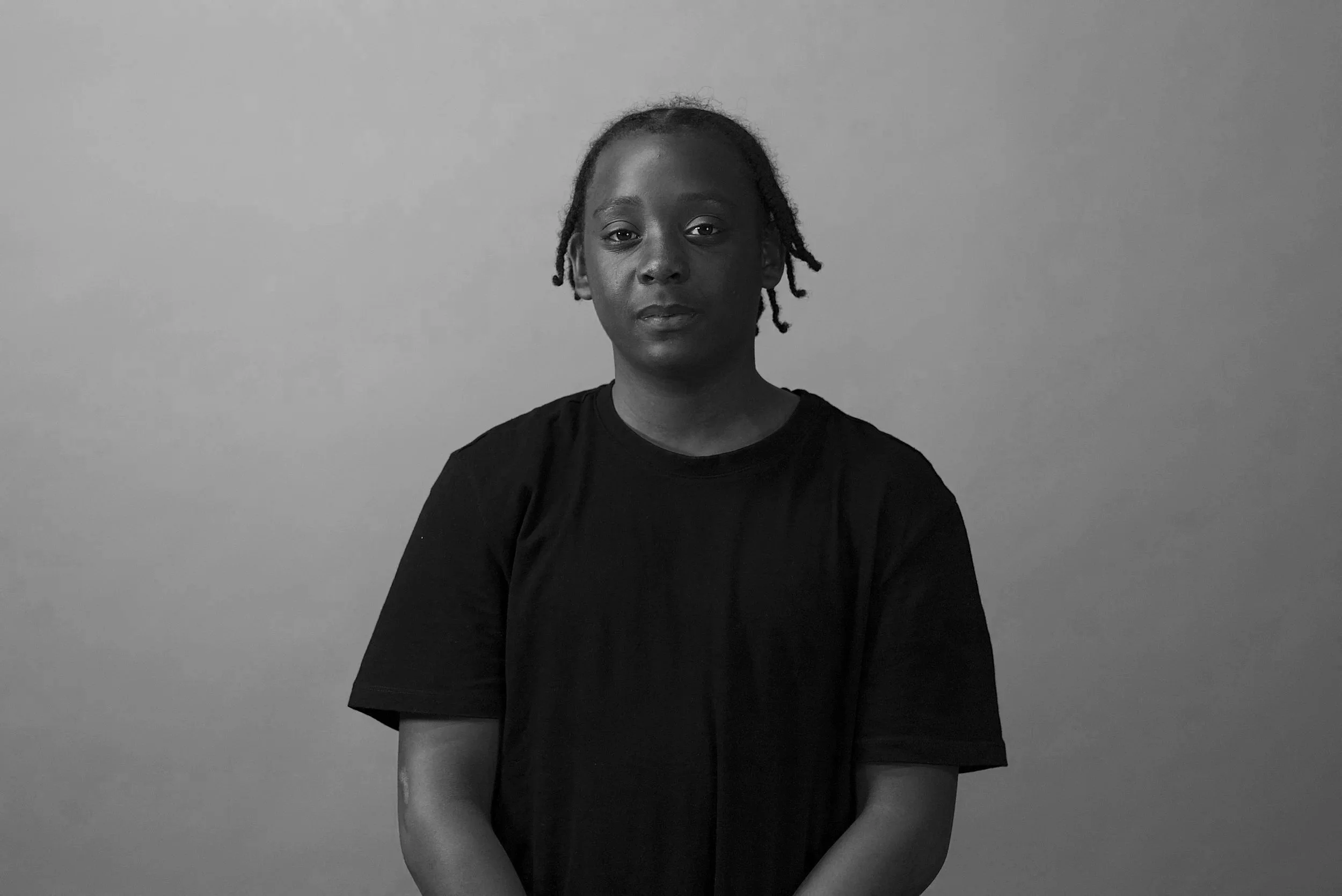 Black and white photo of a young person with short dreadlocks, wearing a black t-shirt, standing against a plain wall.