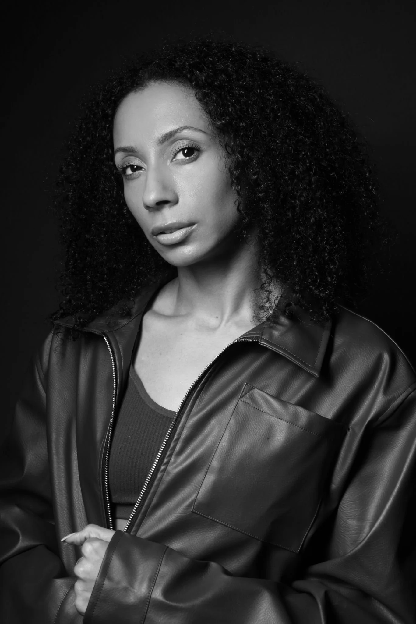 Black and white portrait of a woman with curly hair, wearing a leather jacket and a top, looking directly at the camera with a calm expression.