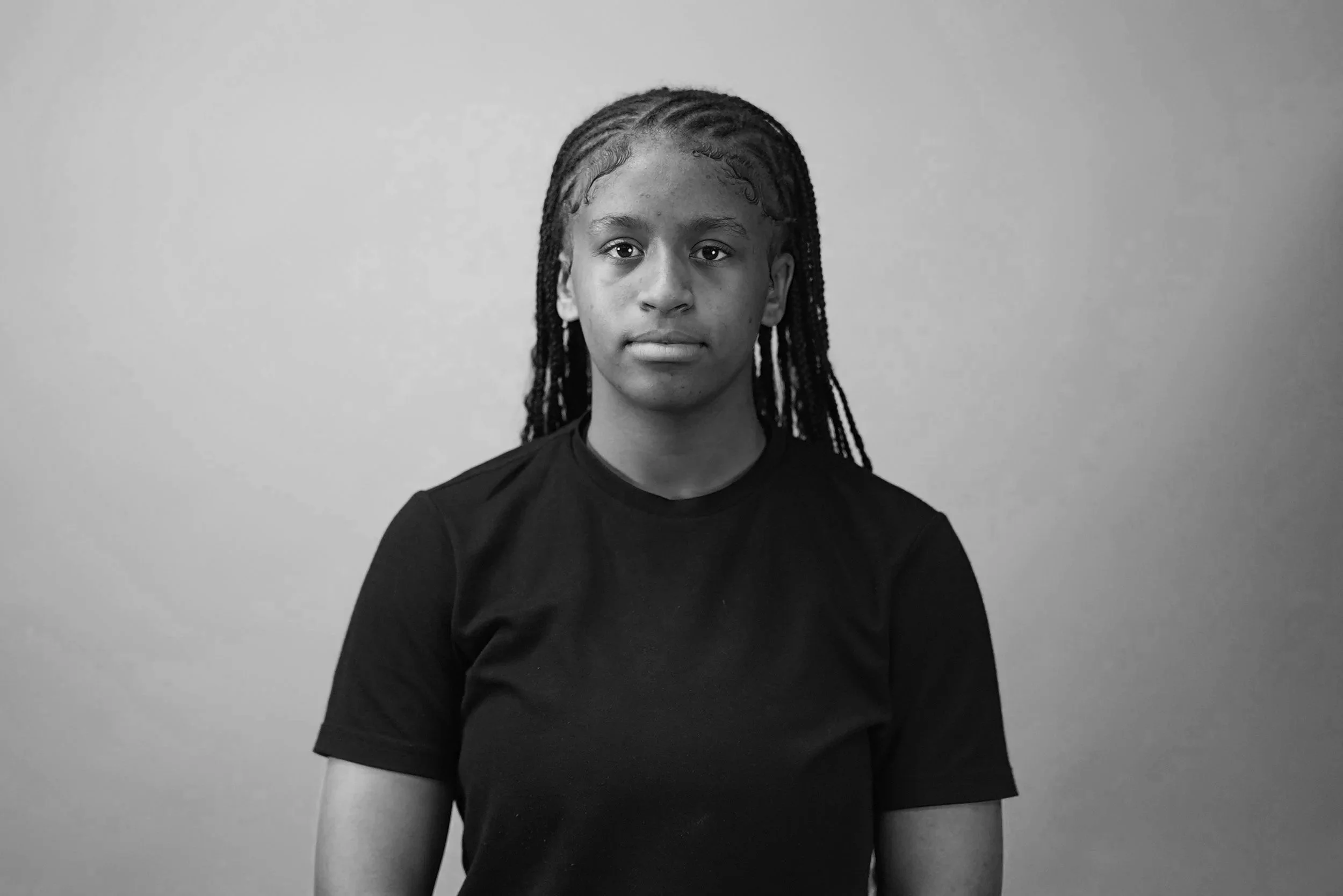 A close-up black and white portrait of a young woman with braids, wearing a dark t-shirt, looking directly at the camera against a plain background.