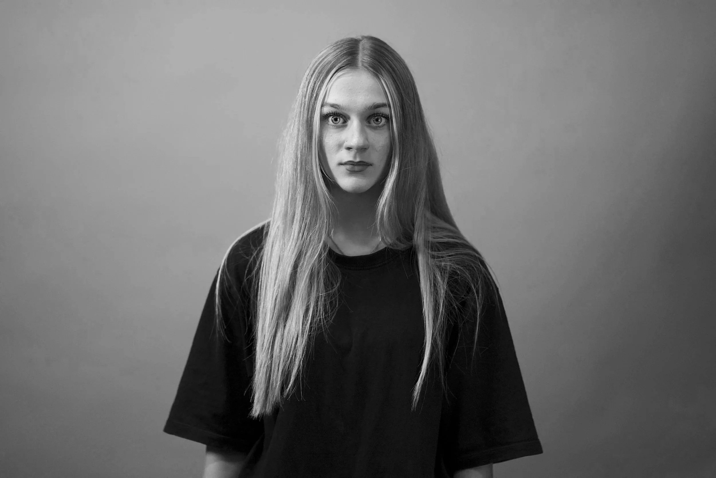 Black and white portrait of a young woman with long straight hair, wide eyes, and wearing a dark long-sleeve shirt, standing against a plain background.