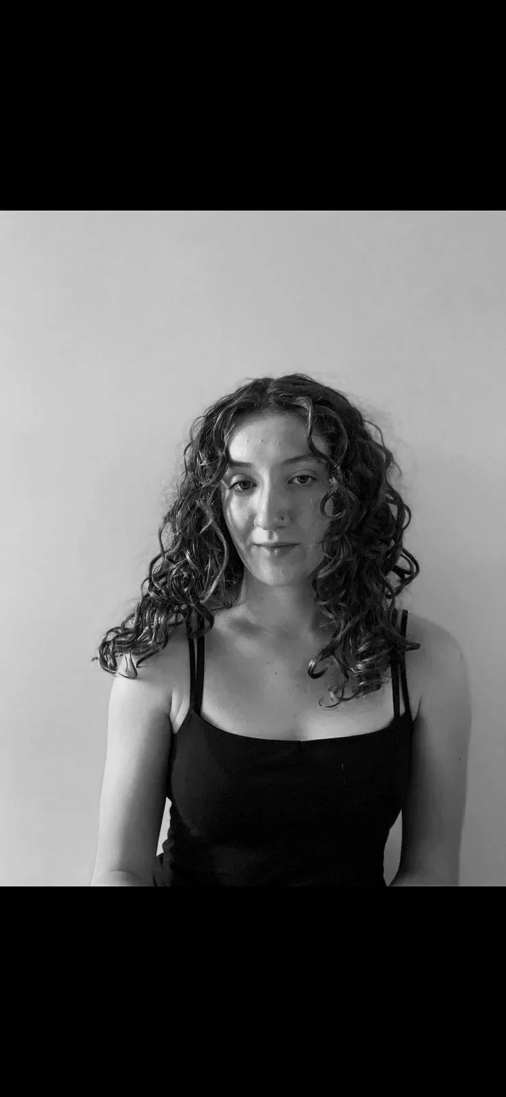 Black and white portrait of a woman with shoulder-length curly hair, wearing a black tank top, standing against a plain light-colored wall.
