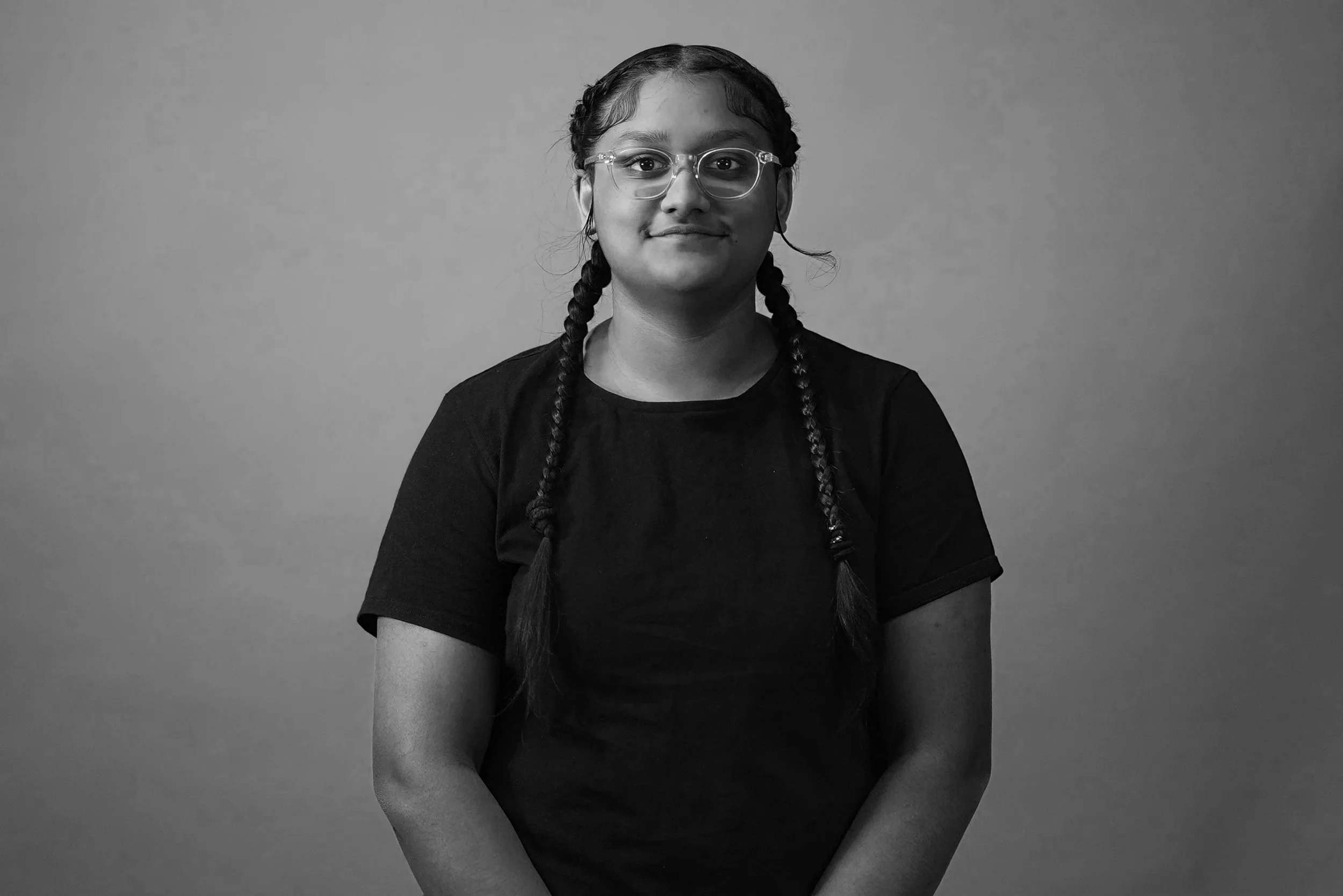Black and white portrait of a woman with glasses, wearing a dark t-shirt, with braided hair, looking directly at the camera against a neutral plain background.