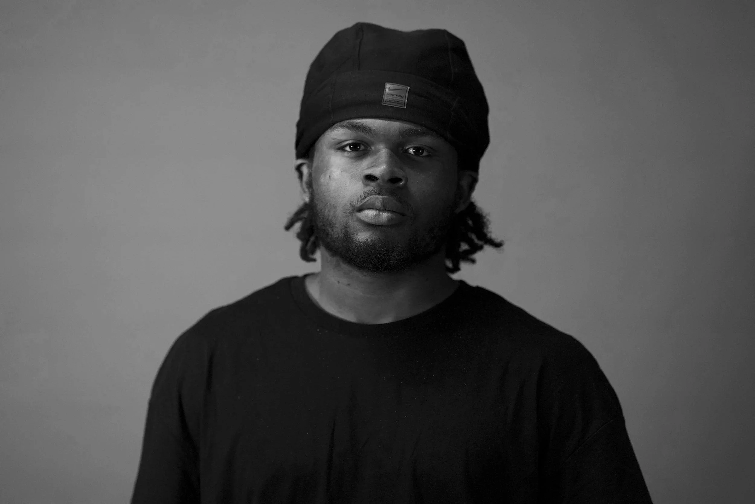 Black and white portrait of a young man wearing a black beanie and a black long-sleeve shirt, looking directly at the camera with a neutral expression.