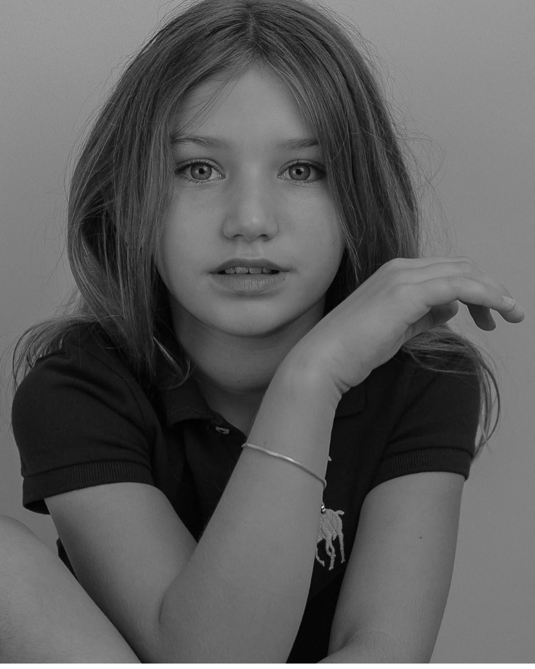 A black and white photo of a young girl with long hair, wearing a polo shirt, looking directly at the camera with her hand resting near her face.