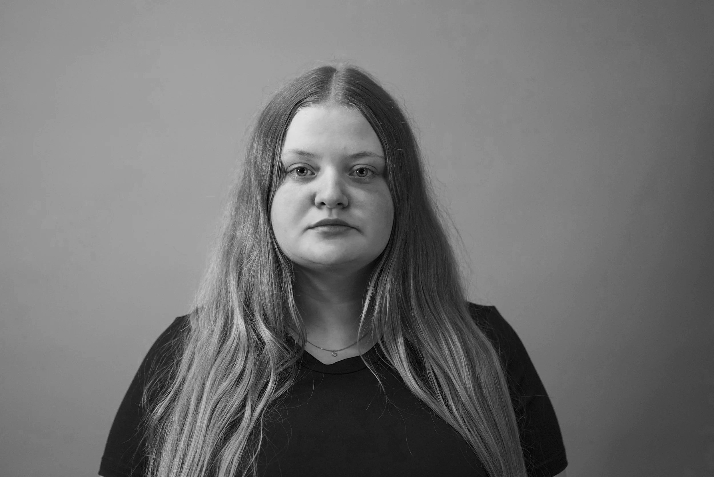 Black and white portrait of a young woman with long hair, wearing a dark top and a necklace, looking directly at the camera with a neutral expression.