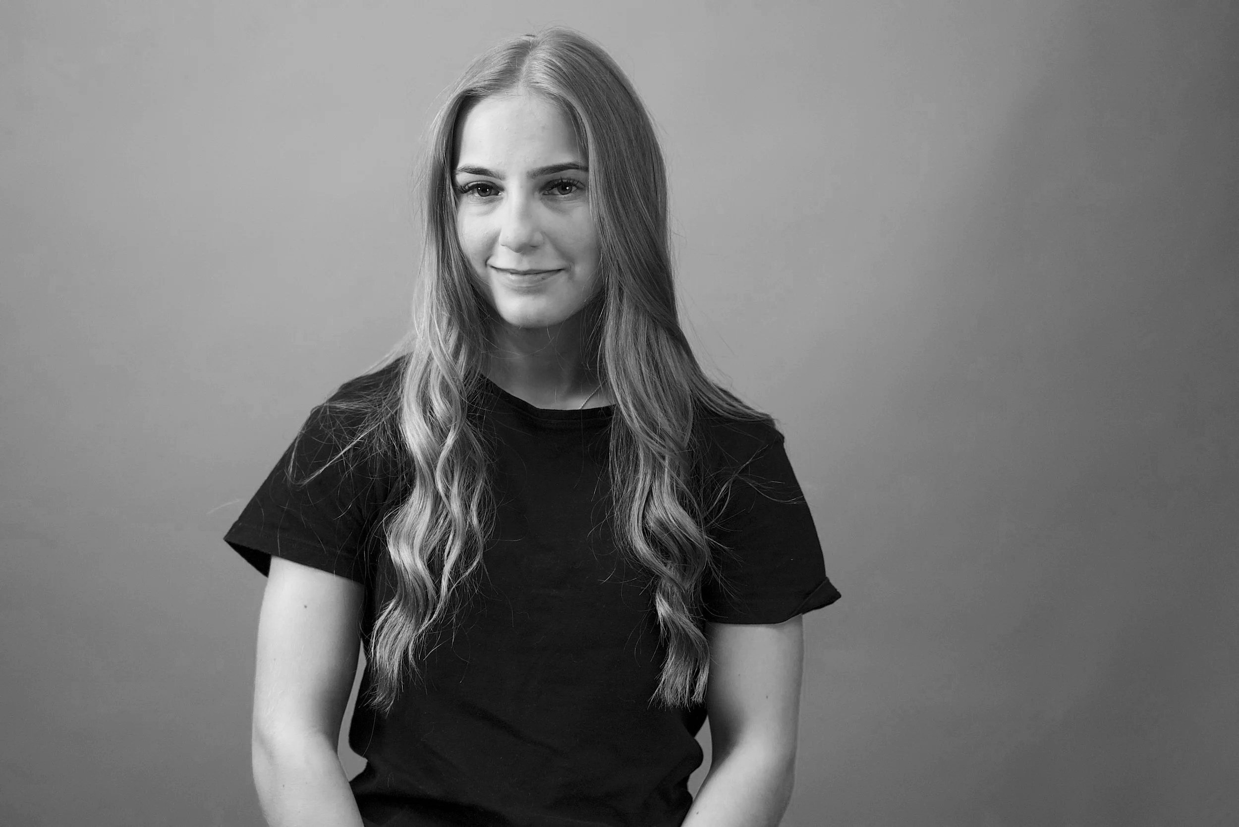Black and white portrait of a young woman with long, wavy hair, smiling slightly, wearing a black T-shirt, against a plain background.