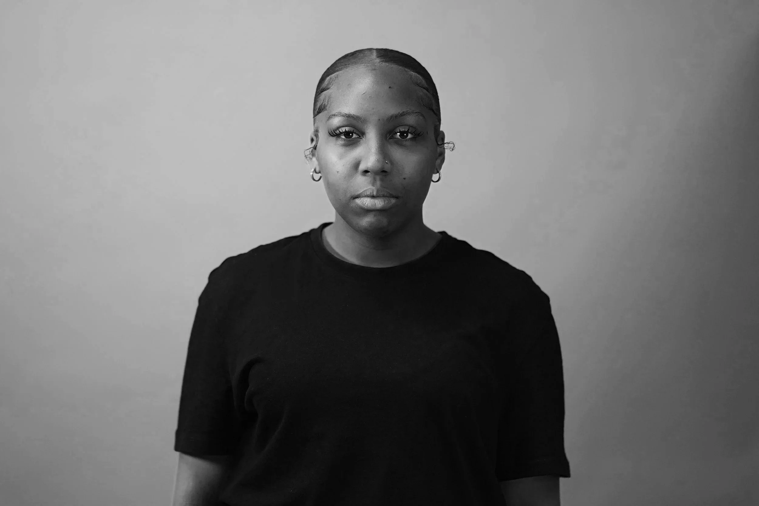A woman with dark skin and short hair styled very close to her scalp, wearing a black shirt, standing against a plain light gray wall, looking straight into the camera with a serious expression.