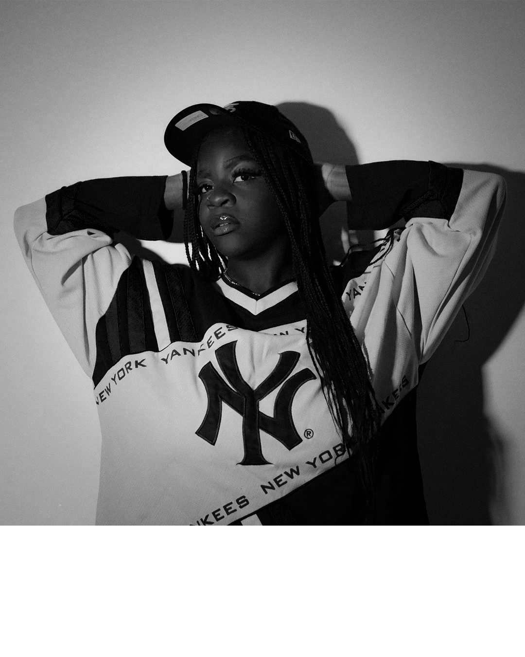 A Black woman wearing a baseball cap backwards, a New York Yankees jersey, and a necklace, posing with her hands behind her head in a black and white photo.