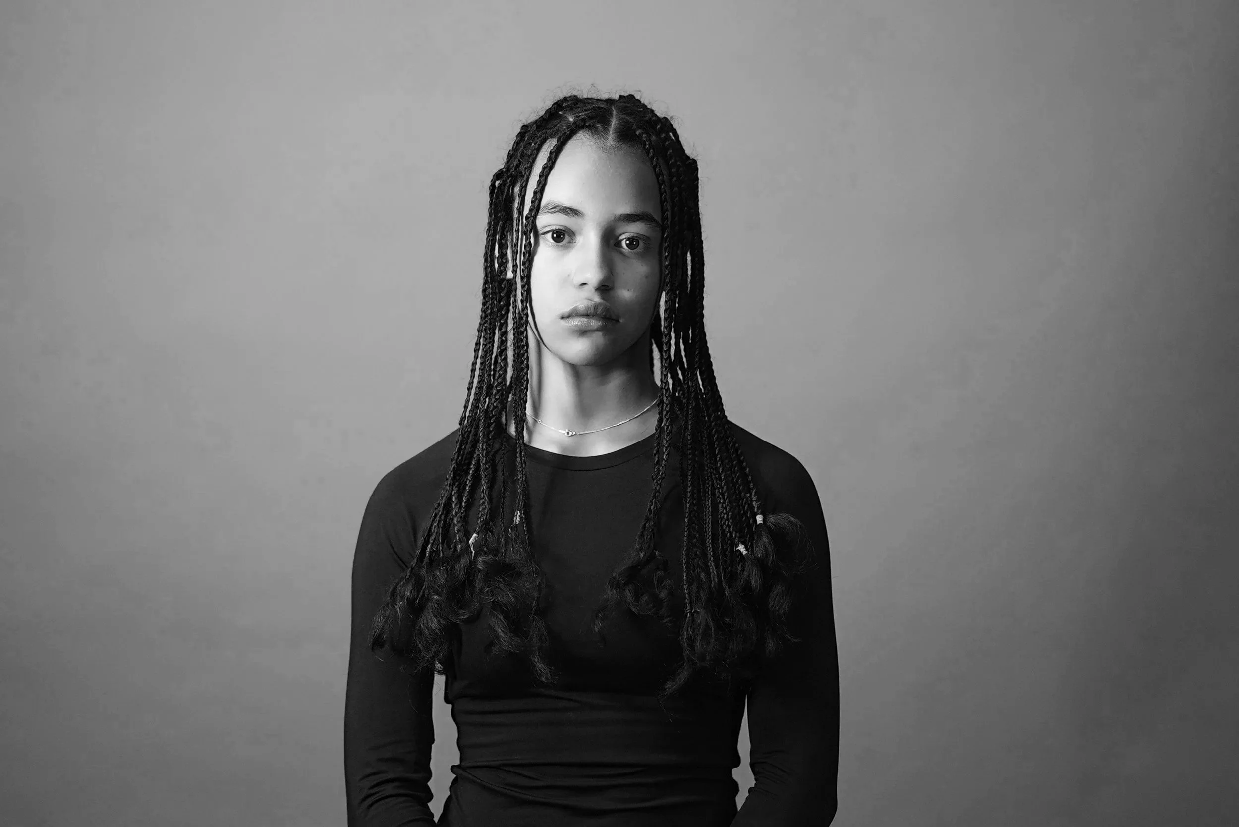 A black and white portrait of a young woman with long braided hair, wearing a dark long-sleeve top, standing against a plain background.