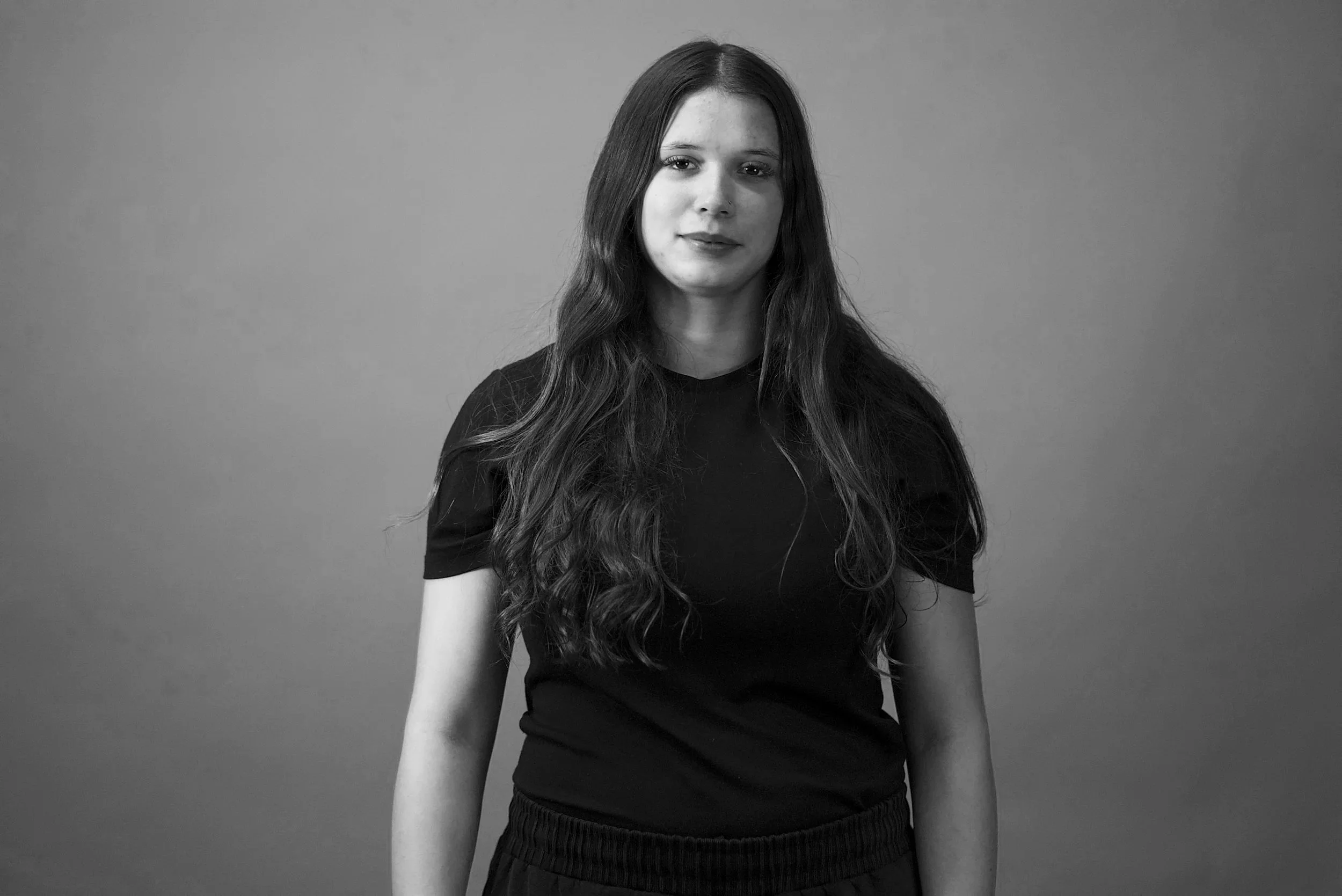 Black-and-white photo of a young woman with long, wavy hair, wearing a black shirt, standing against a plain background.