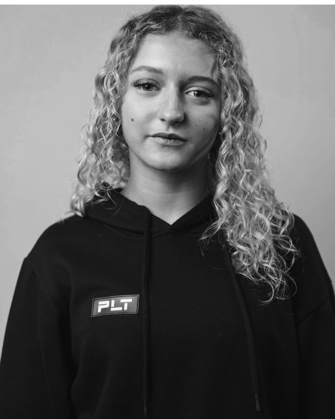 A black-and-white portrait of a woman with curly shoulder-length hair, wearing a black hoodie with a PLT logo on the chest, standing against a plain background.