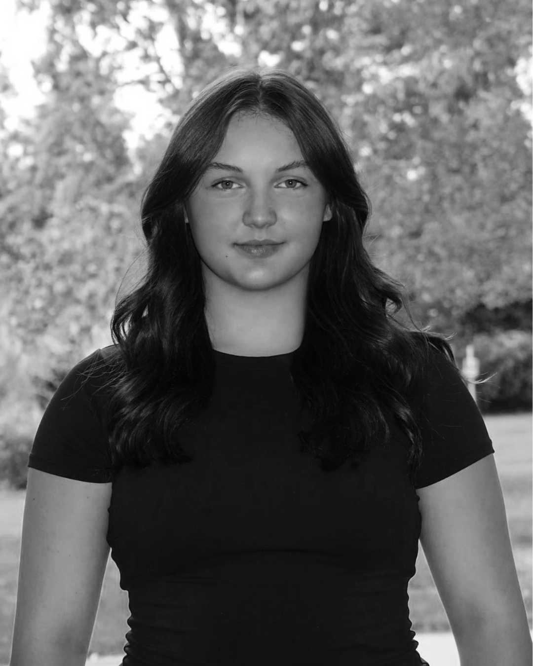 Black-and-white portrait of a young woman with long dark hair, wearing a black t-shirt, standing outdoors with trees in the background.