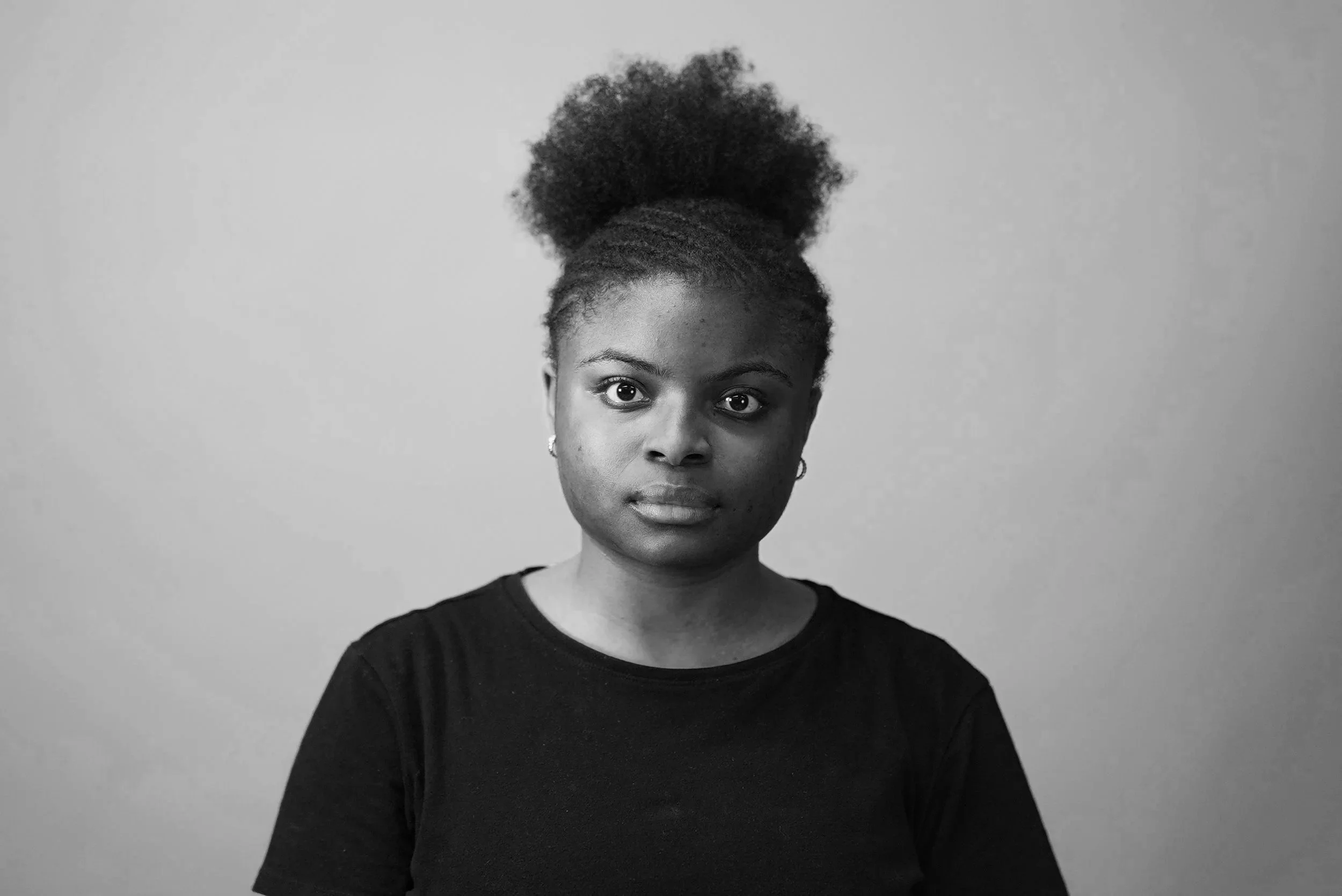 A black-and-white portrait of a woman with natural hair styled in a high puff, looking directly at the camera against a plain background, wearing a dark top.