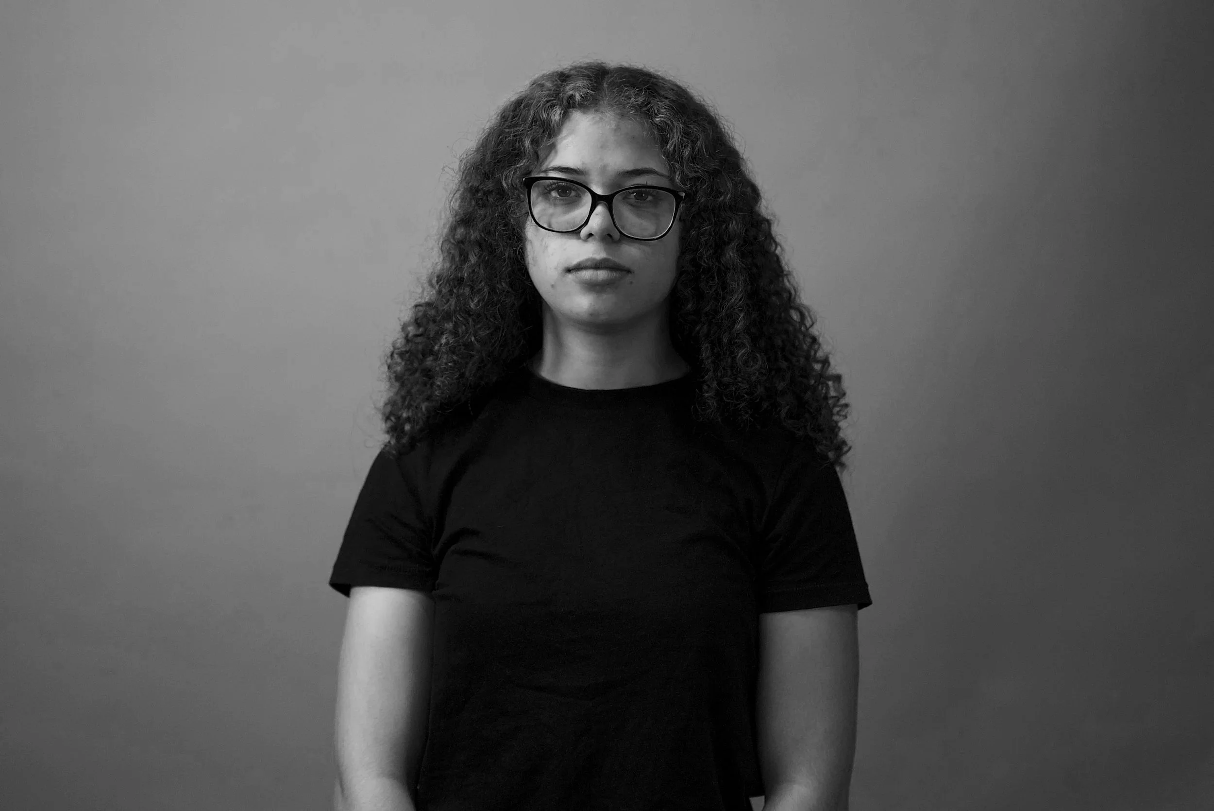 Black and white portrait of a woman with curly hair, wearing glasses and a black shirt, standing against a plain background.