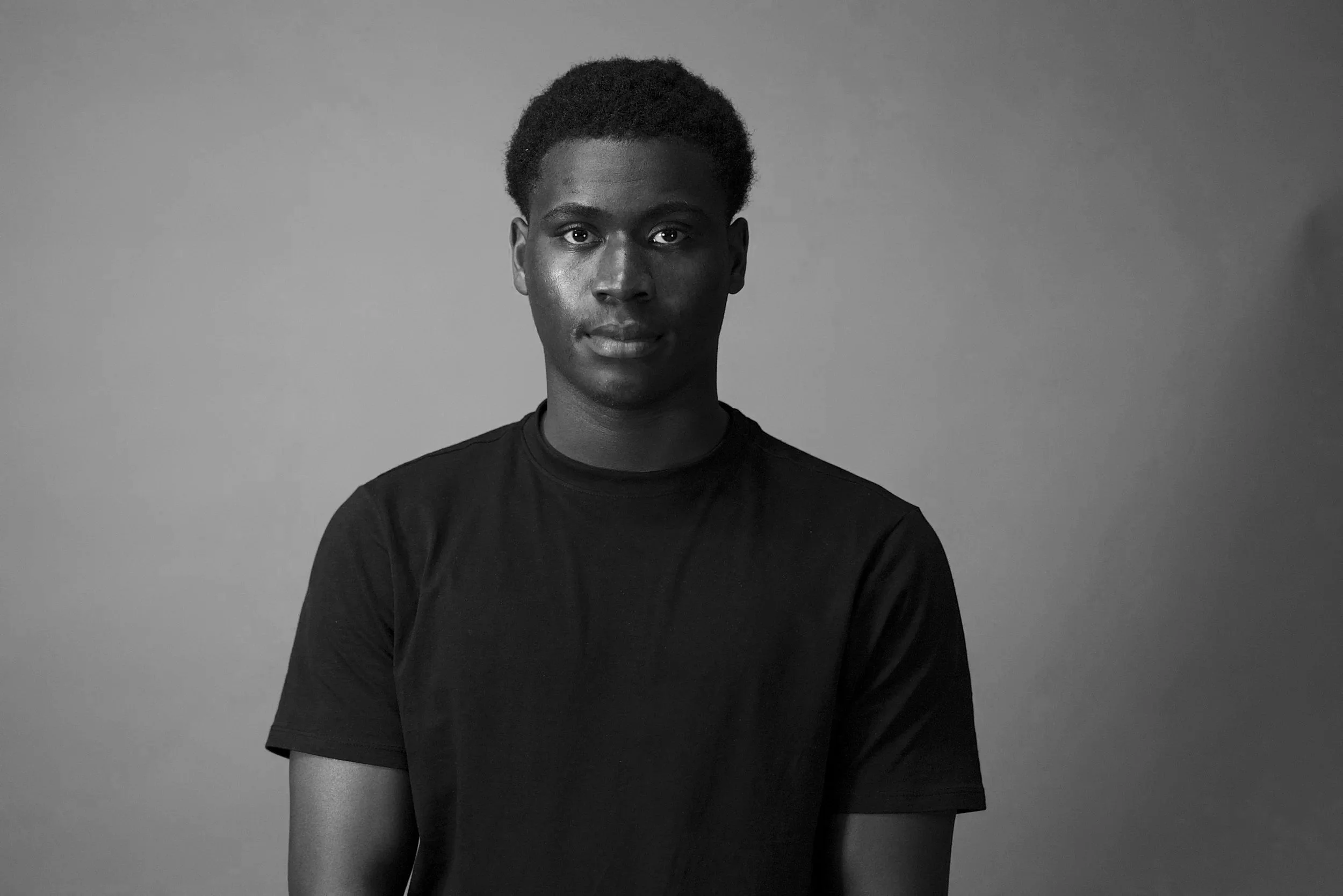 A young man with dark skin and short curly hair, wearing a black t-shirt, standing against a plain gray background.