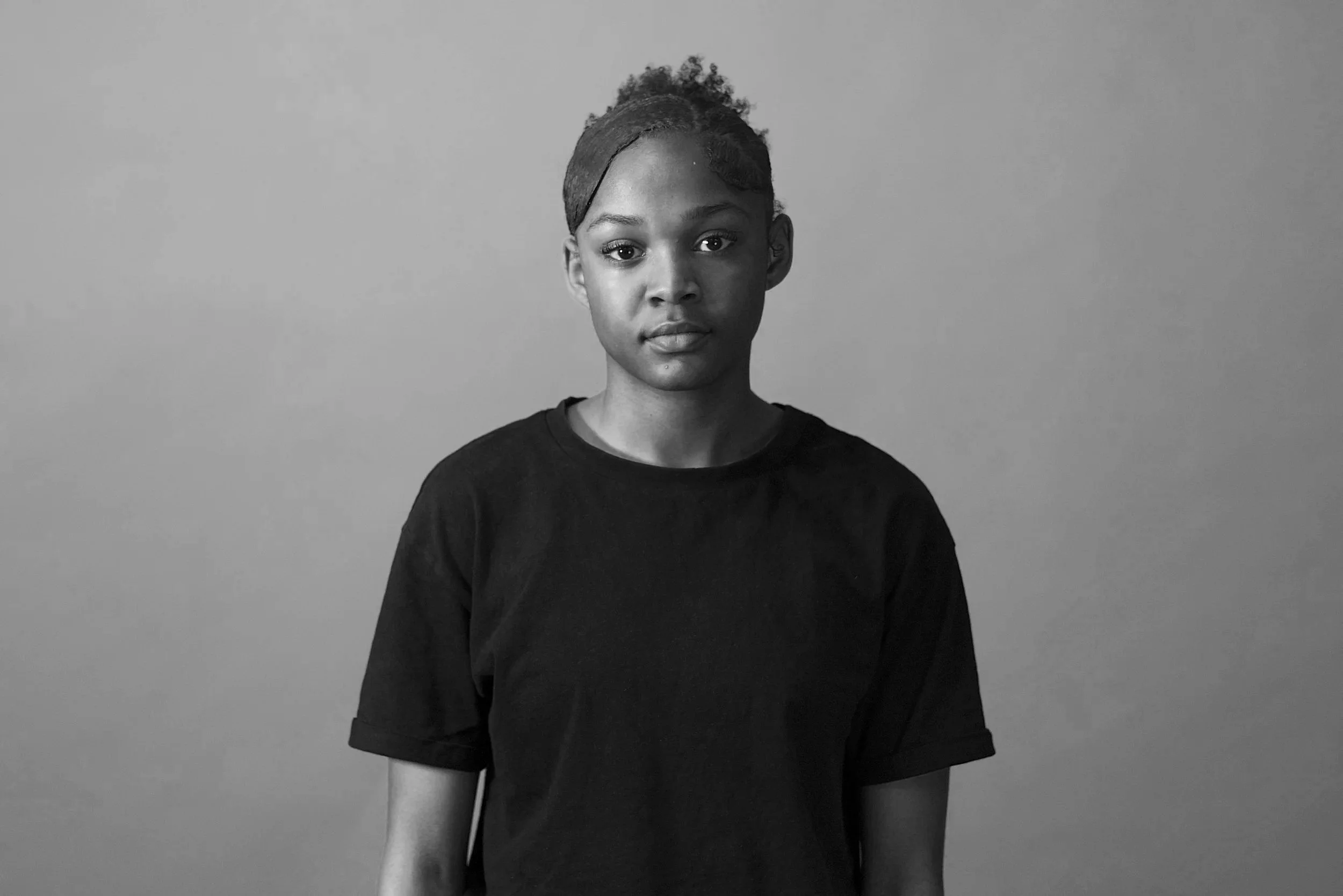 A black and white portrait of a young woman with short, curly hair tied back with a headband, wearing a dark T-shirt, standing against a plain background.