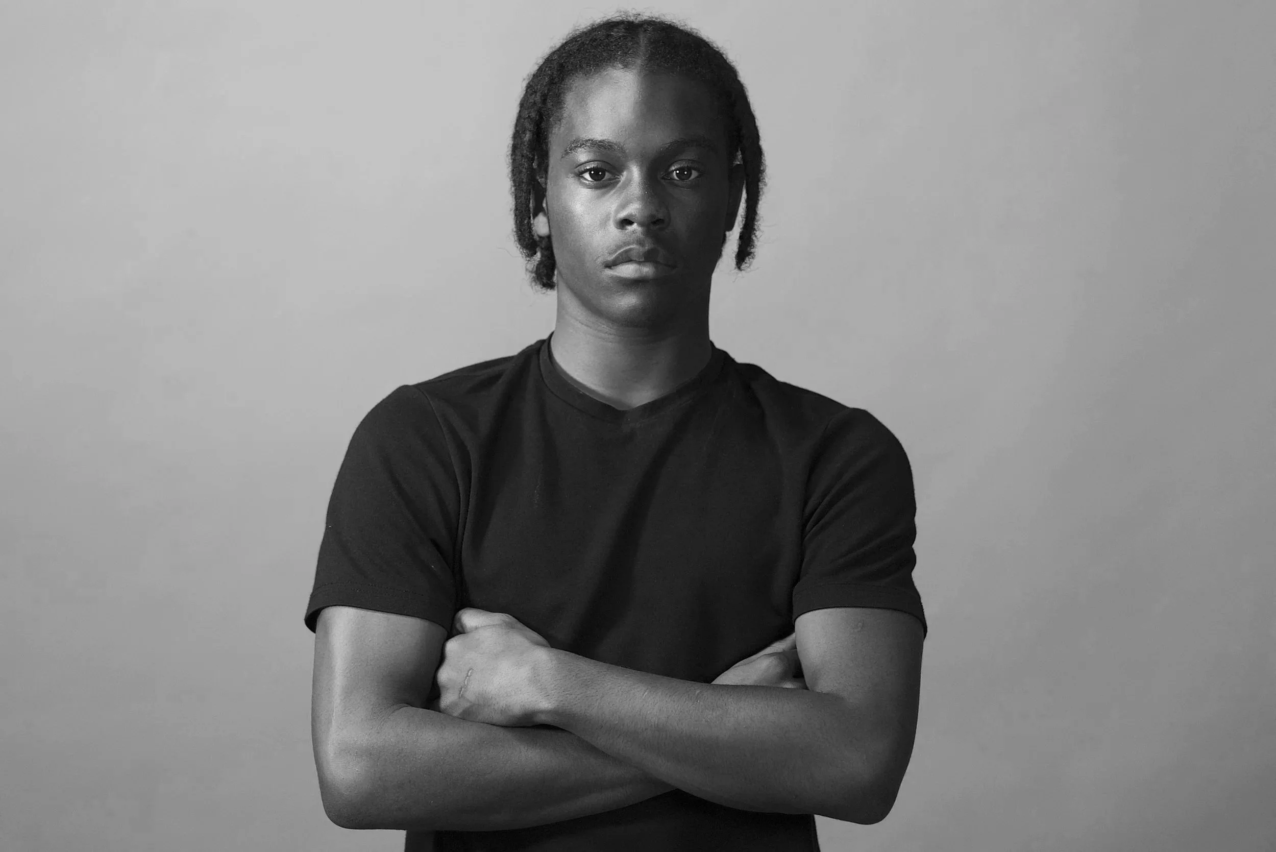 Black and white photo of a woman with short dreadlocks, wearing a black t-shirt, crossing her arms, looking serious against a plain background.