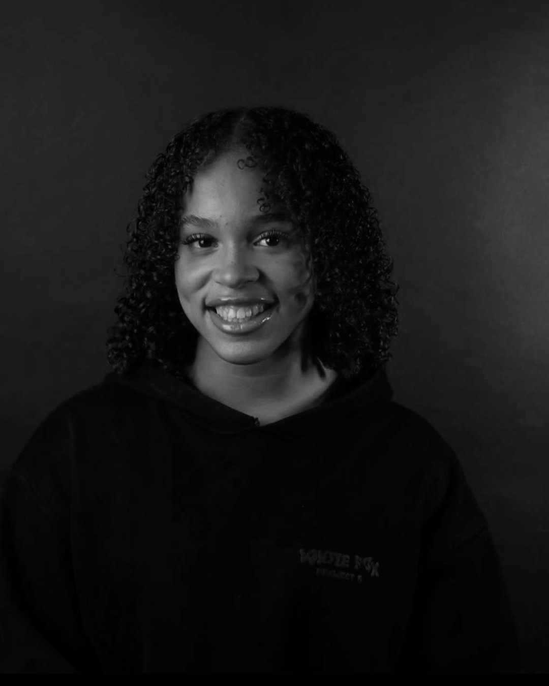 Black and white portrait of a young woman smiling, with curly hair, wearing a dark hoodie, against a dark background.