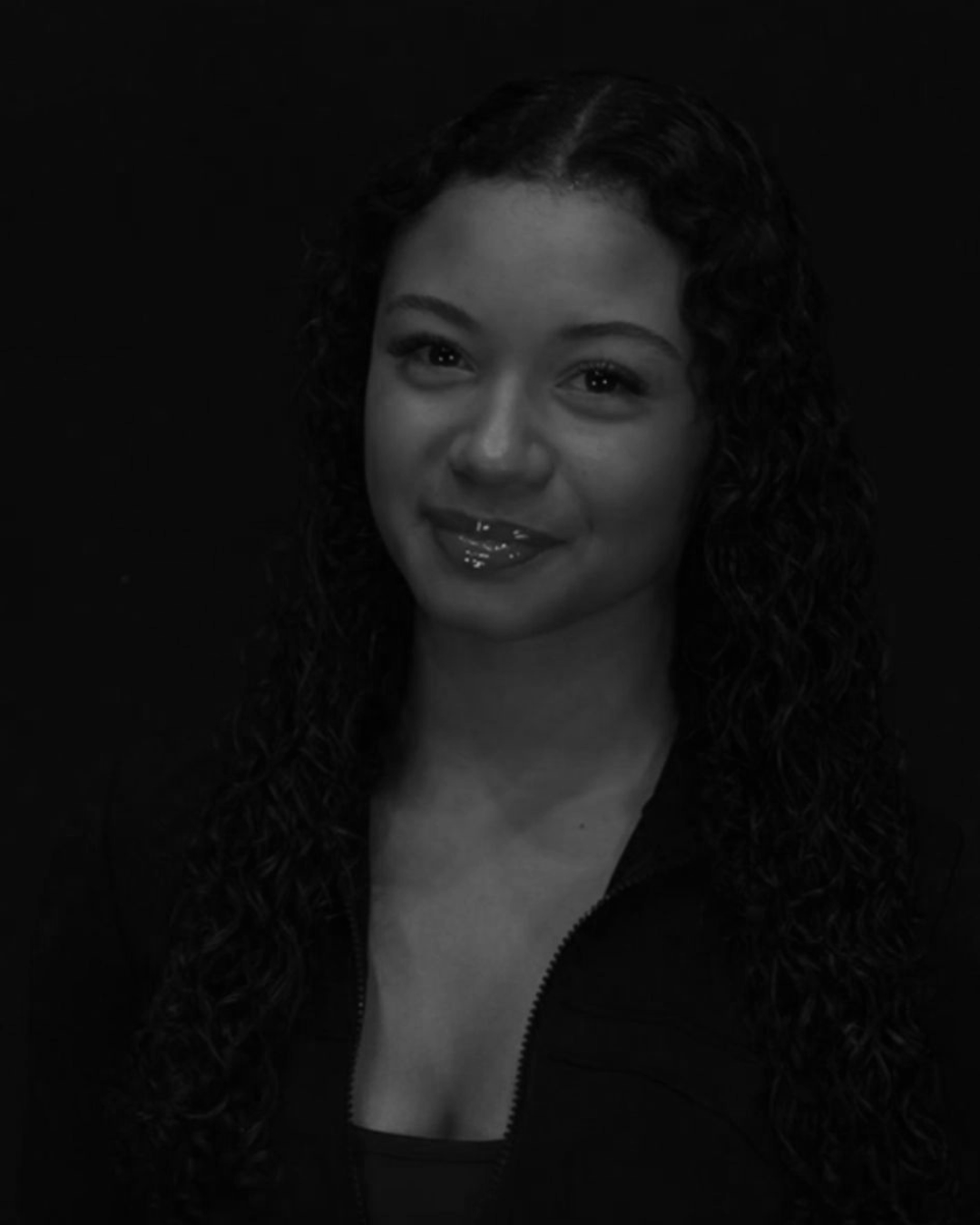 A woman with curly hair and a slight smile, wearing a dark top, against a dark background.