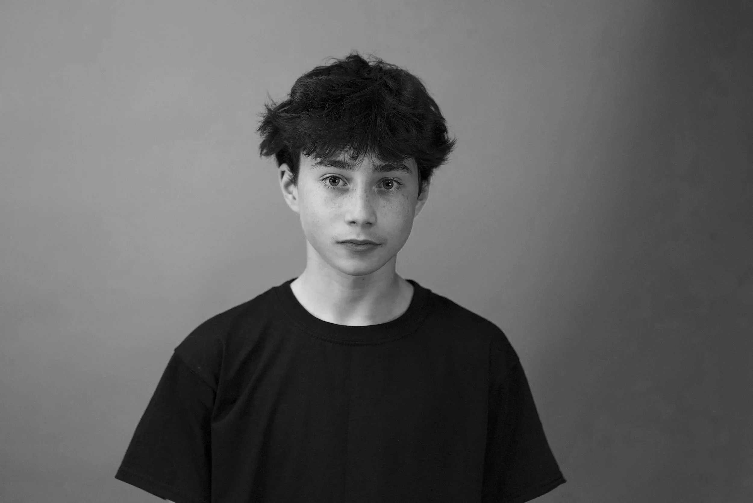 A black and white photo of a young person with short, tousled hair wearing a dark shirt, standing against a plain background.