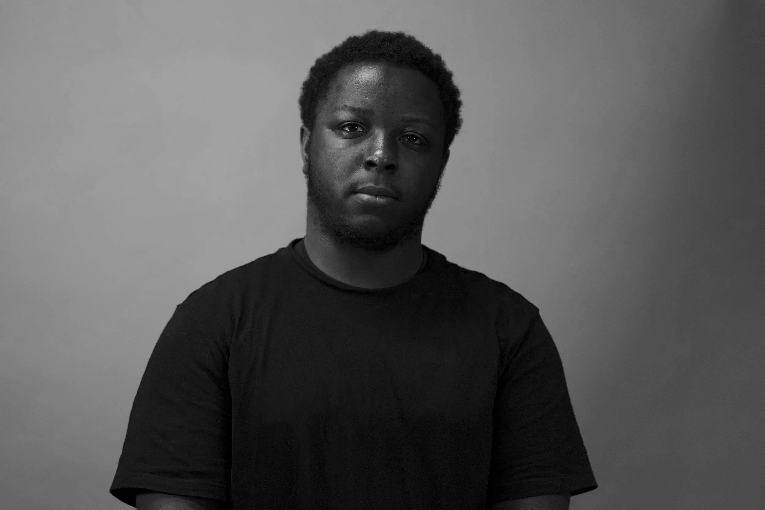 A portrait of a man with dark skin and short curly hair, wearing a black T-shirt, against a plain light grey background.