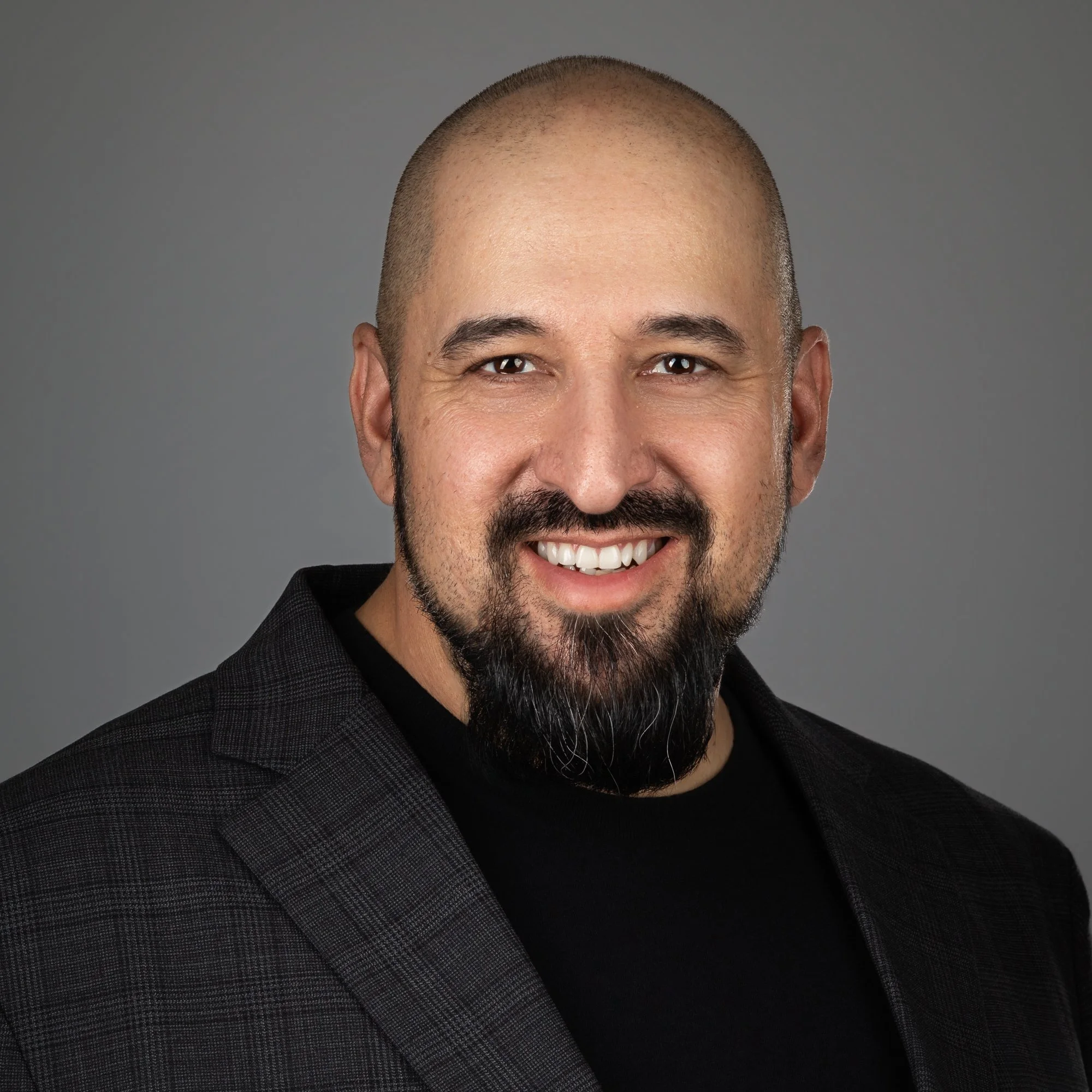 Close-up of a man with a bald head, dark beard, smiling, wearing a dark blazer, against a gray background.