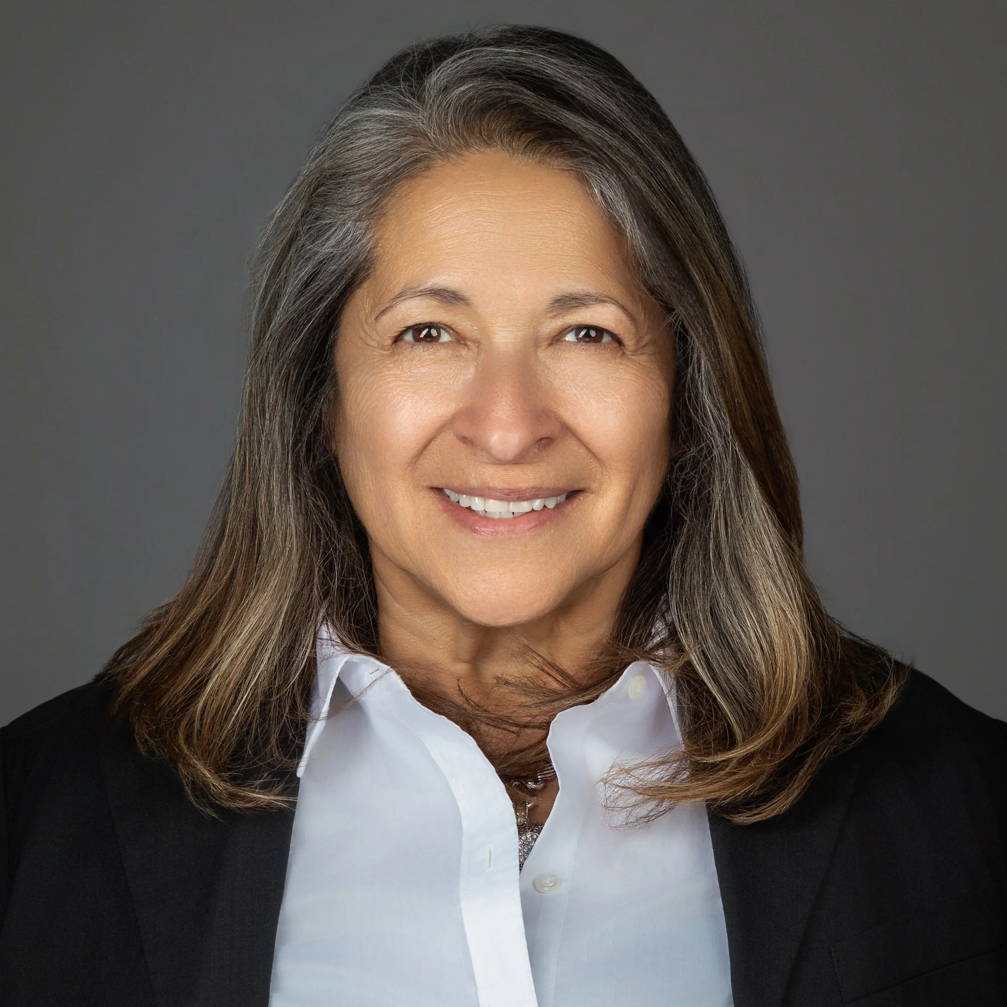 Close-up of a middle-aged woman with long brown and gray hair, smiling and wearing a white shirt and black blazer against a gray background.