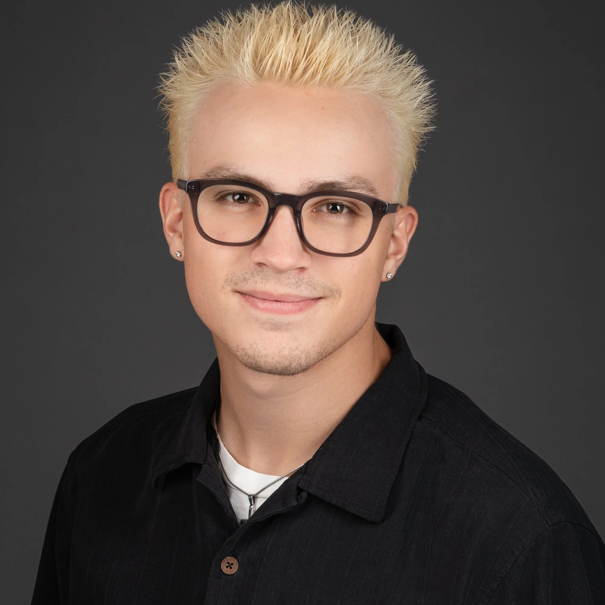 Close-up portrait of a young man with blonde hair styled upwards, wearing dark rectangular glasses, a black shirt, and a silver necklace, against a dark background.