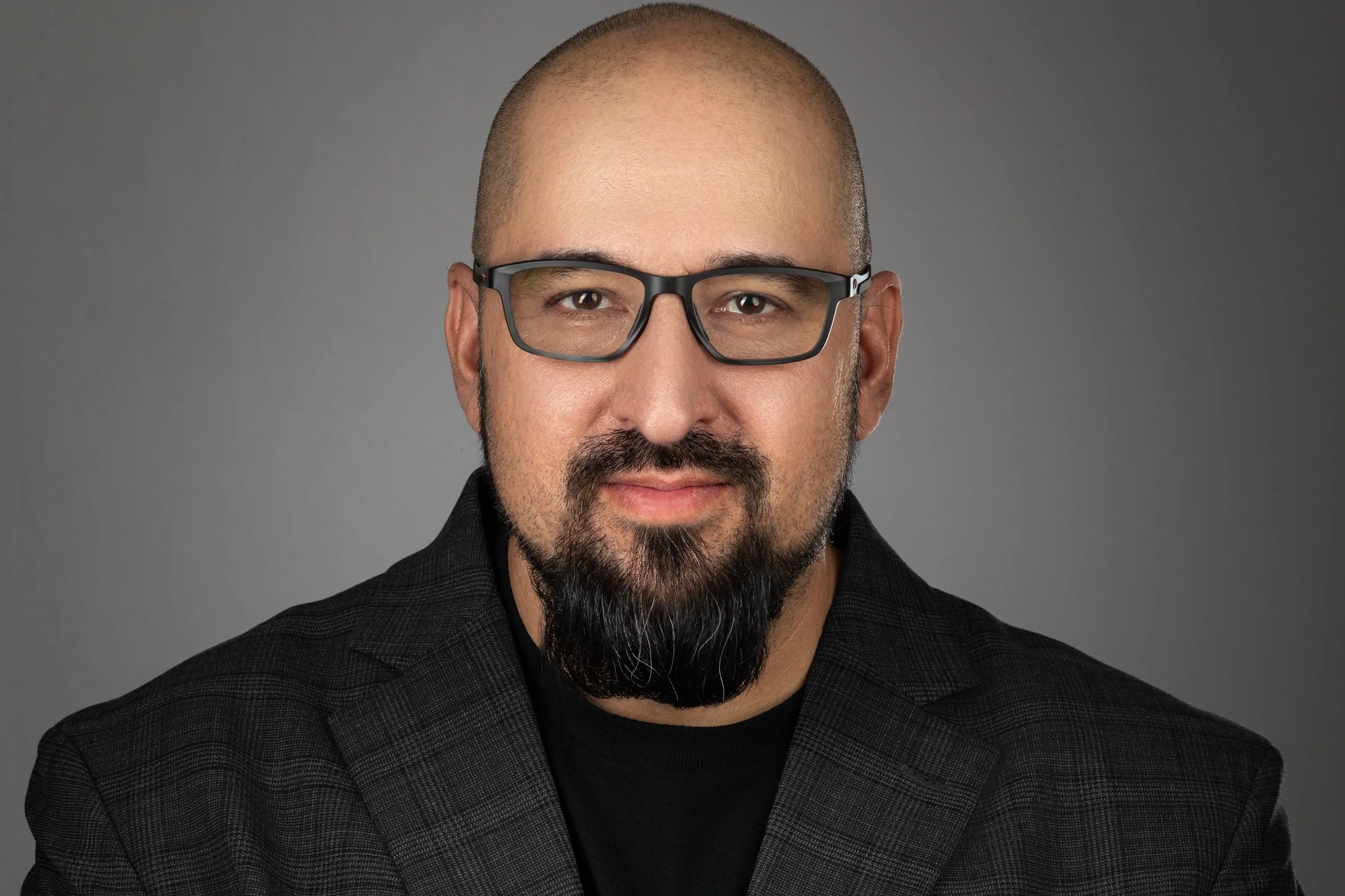 A close-up portrait of a man with glasses, a shaved head, a goatee, and a mustache, wearing a dark checkered blazer and black shirt, against a plain gray background.